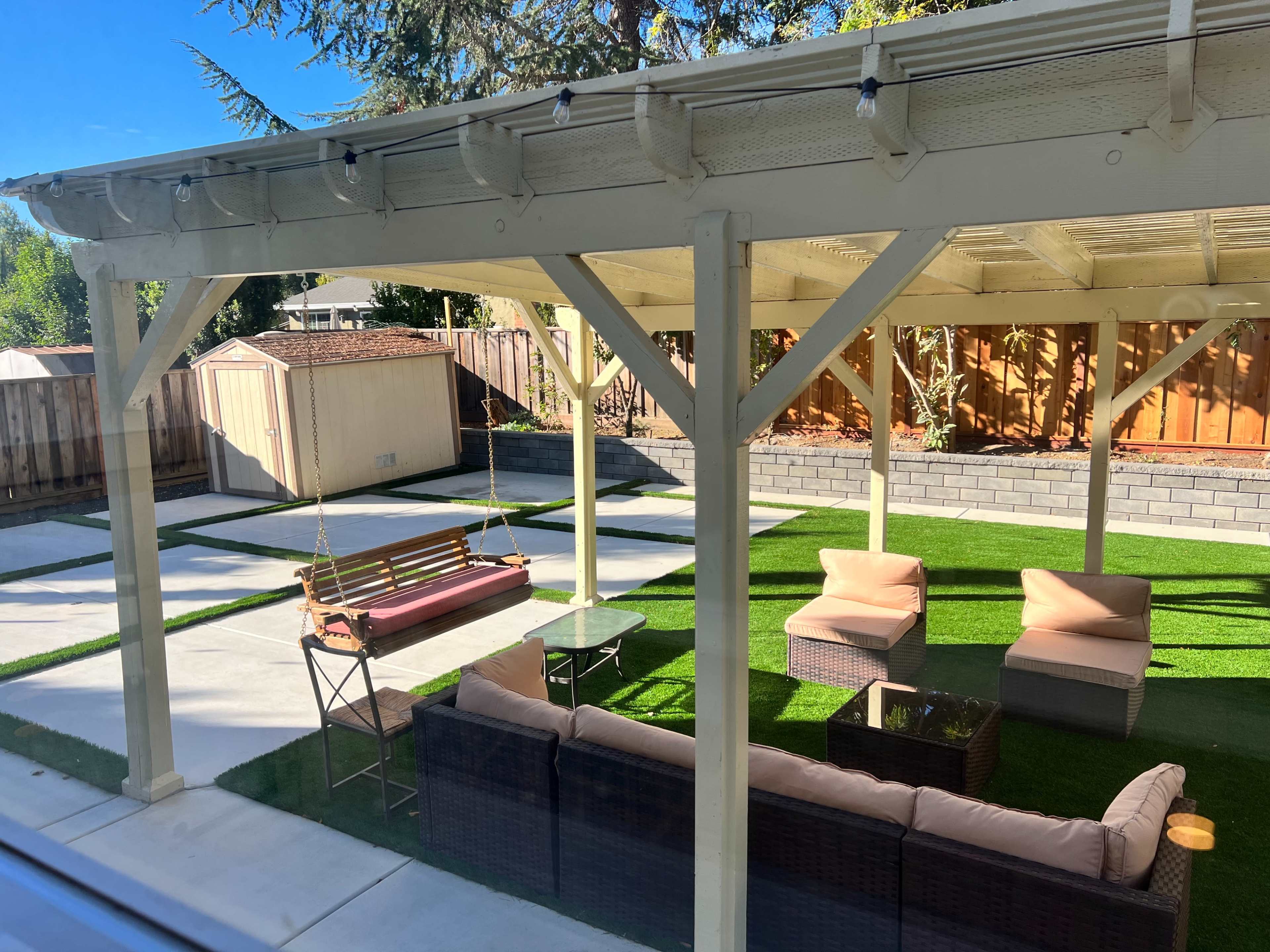 The image shows a backyard patio with a covered seating area, including a couch and chairs, a swing, and a small table, all situated on artificial grass.