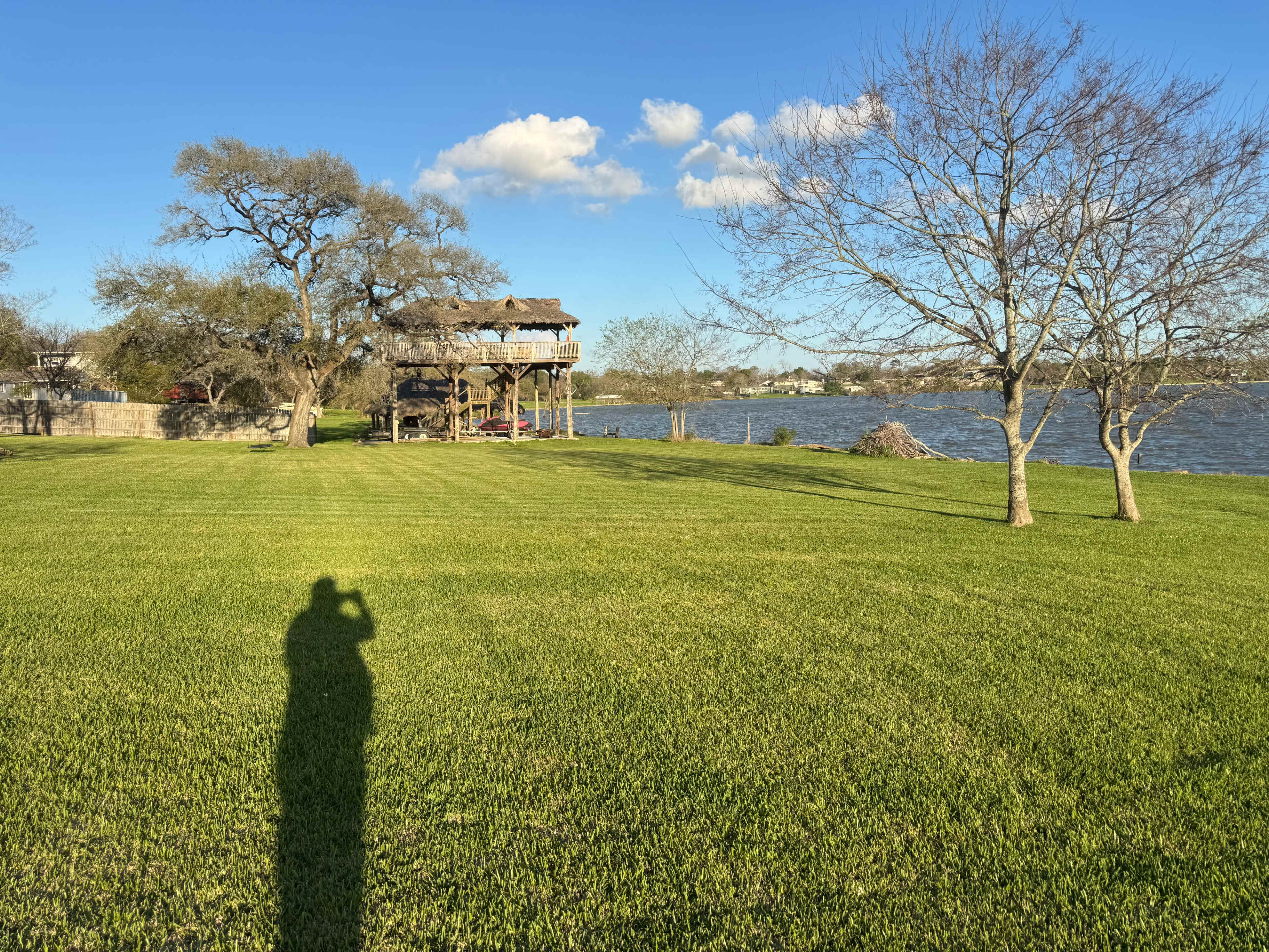 A grassy area extends toward a river, with a large wooden structure and several trees in the background under a blue sky.