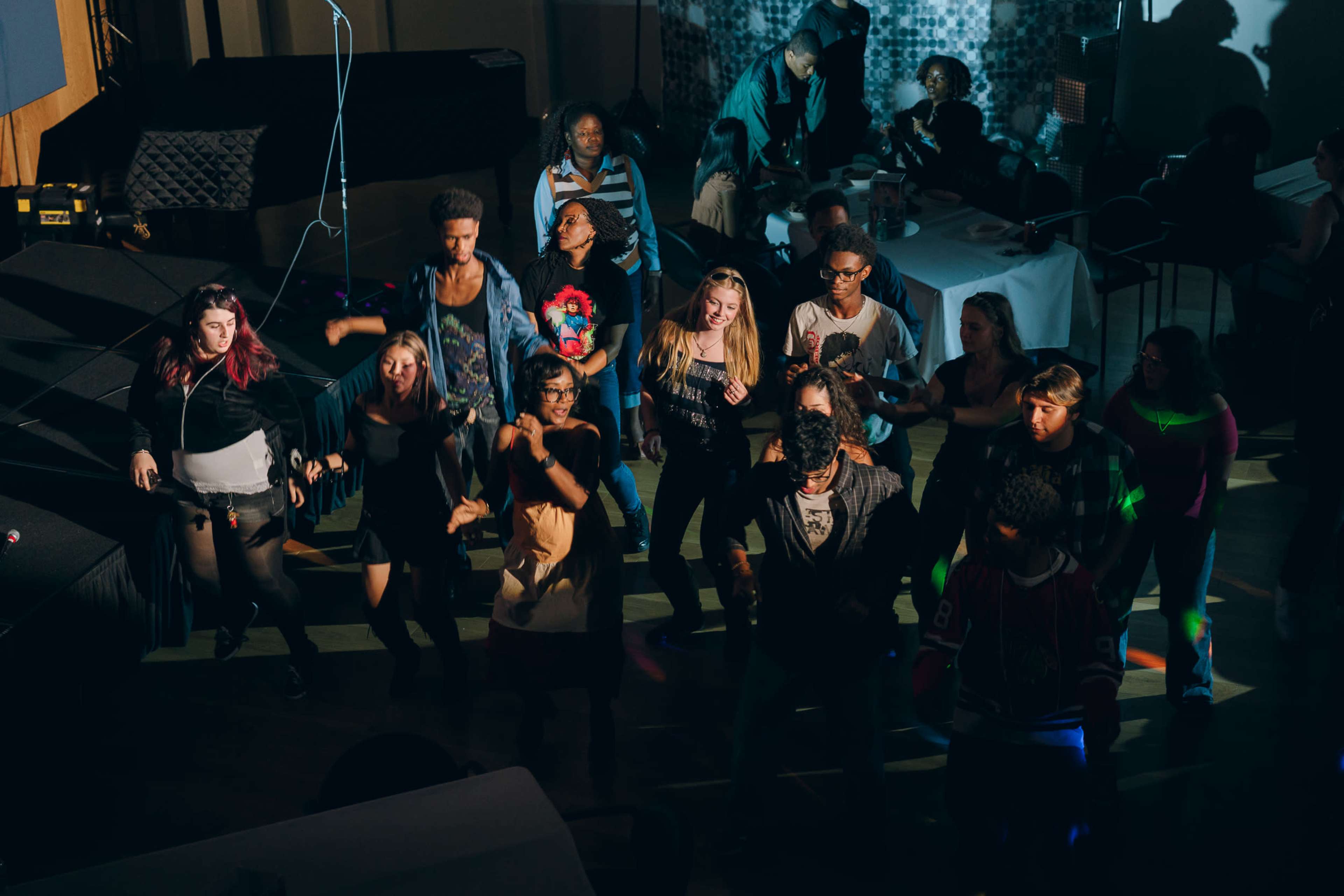 A group of diverse individuals is dancing together in a brightly lit venue while others socialize in the background.