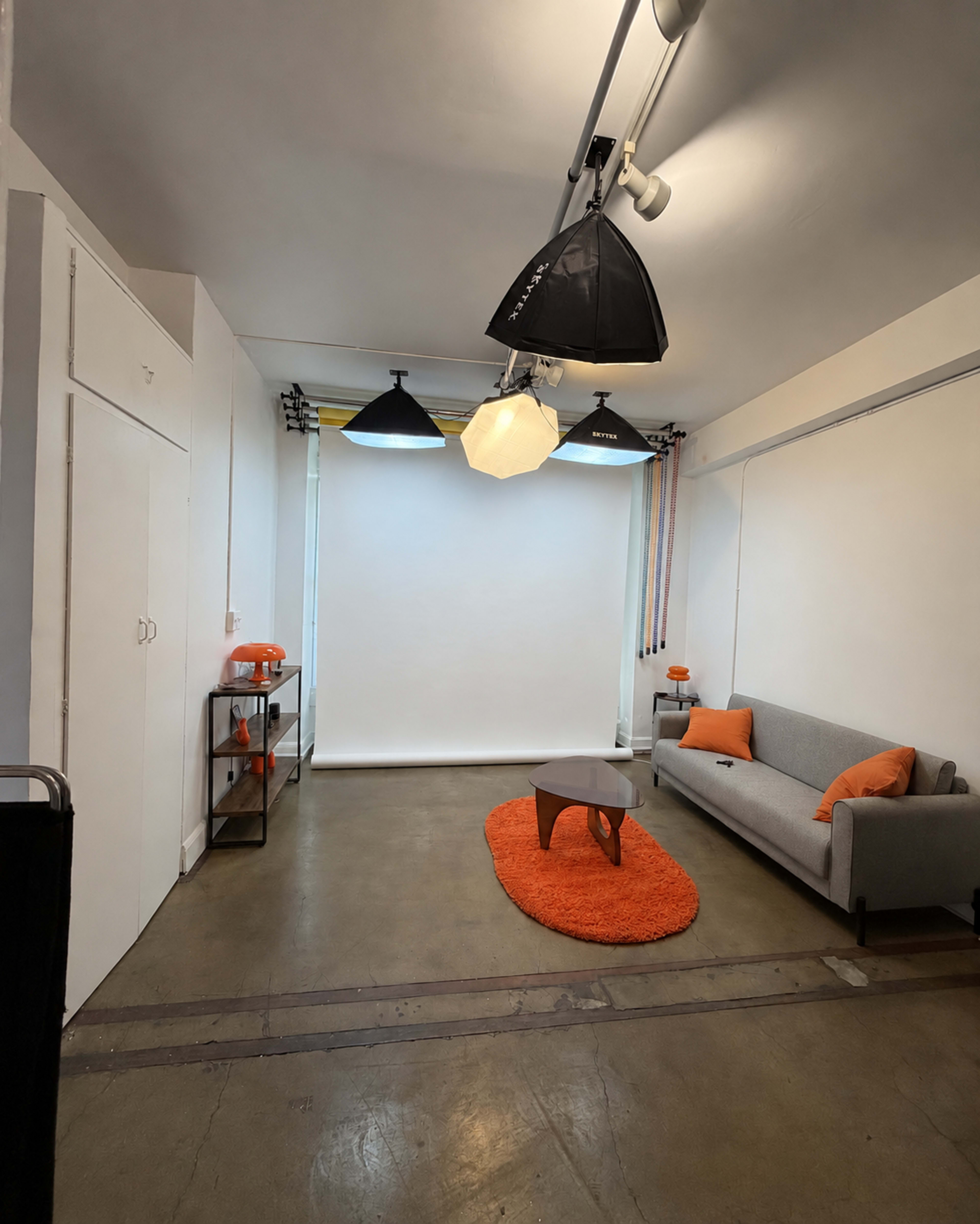 The image depicts a minimalistic interior studio space featuring a gray sofa, an orange rug, and various lighting equipment suspended from the ceiling.