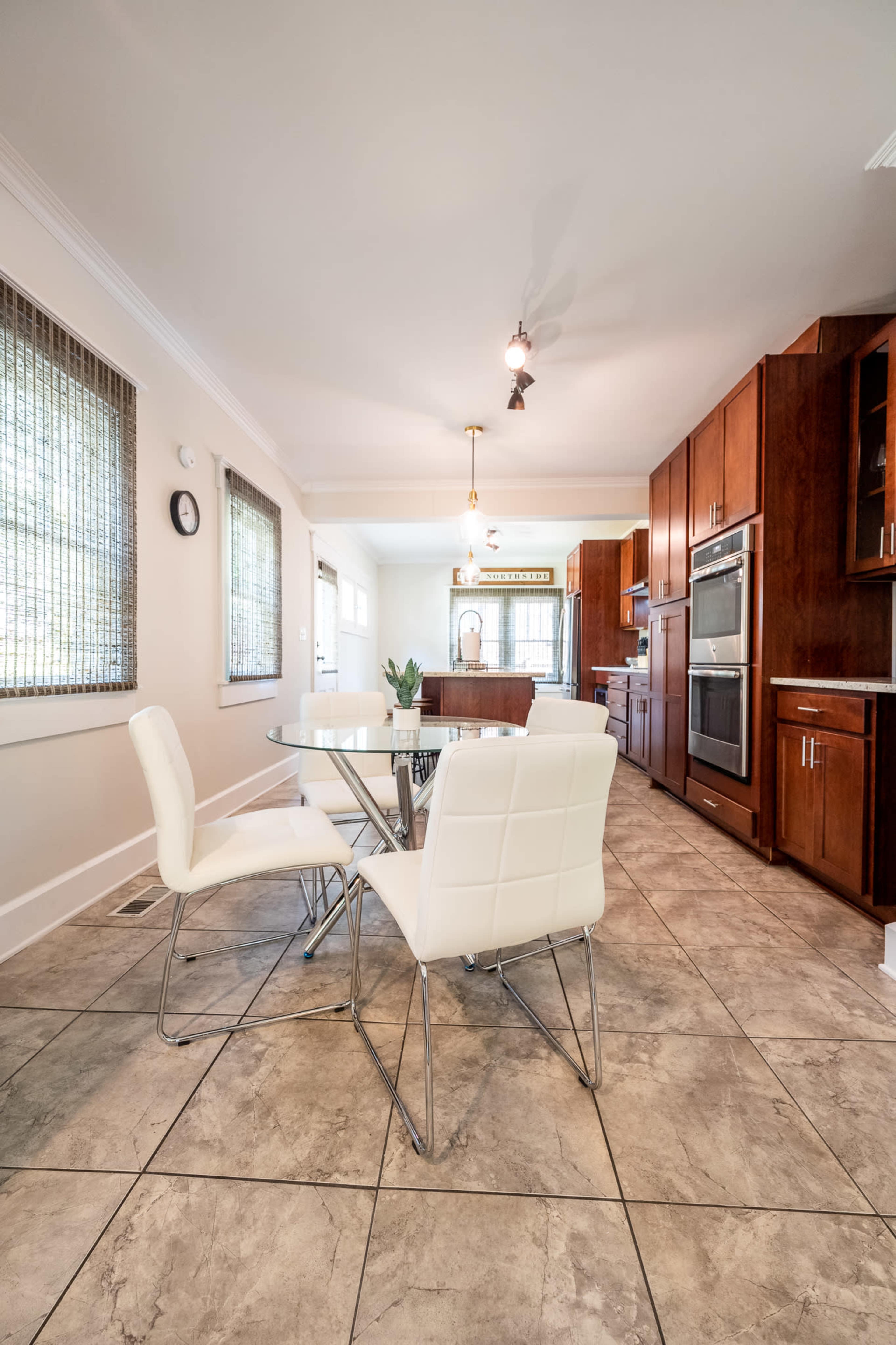 A modern kitchen features a glass dining table with white chairs, complemented by wood cabinets and tiled flooring.
