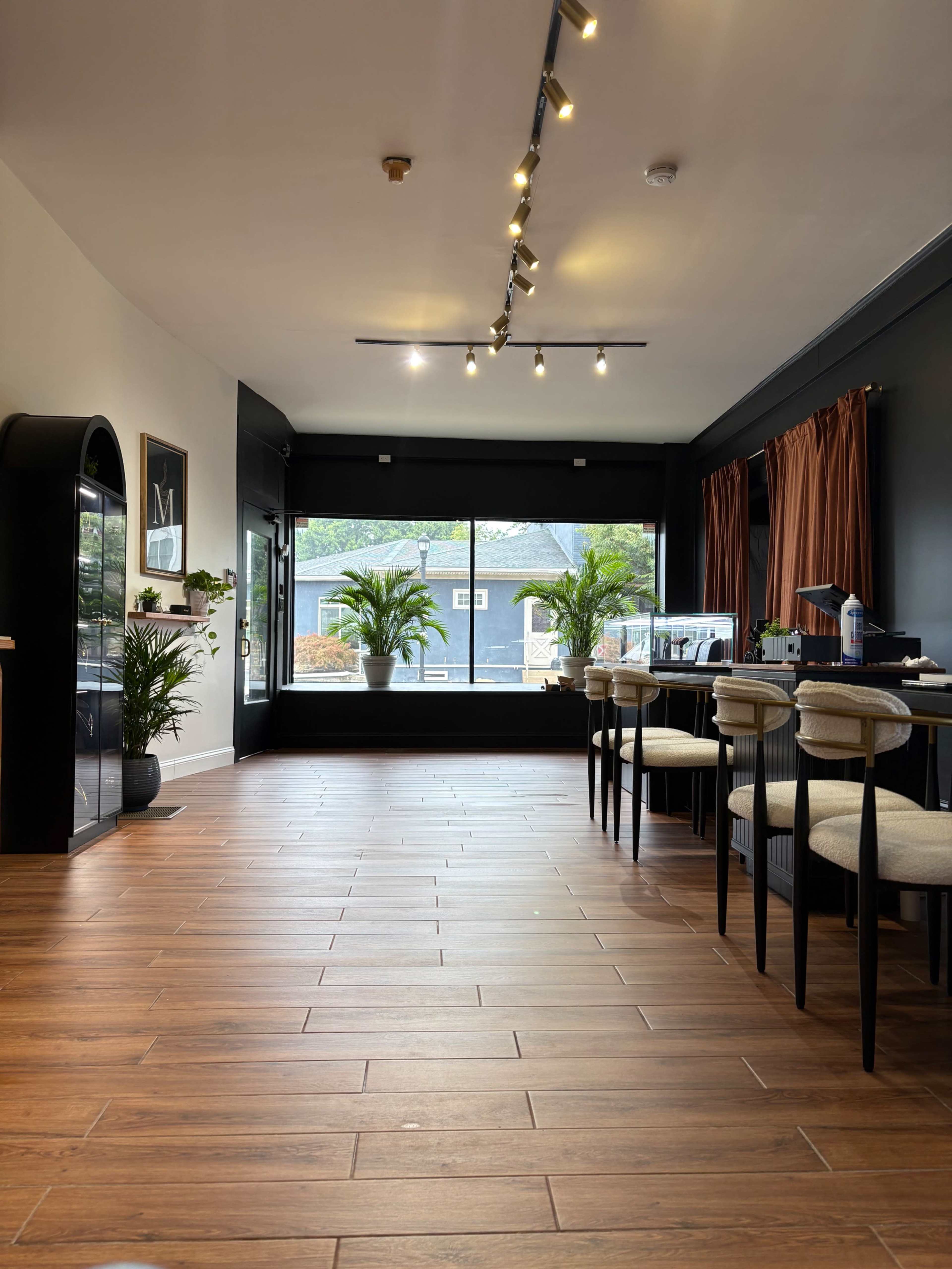 The image shows a well-lit interior of a modern space with wooden flooring, seating along one side, and large windows featuring potted plants.
