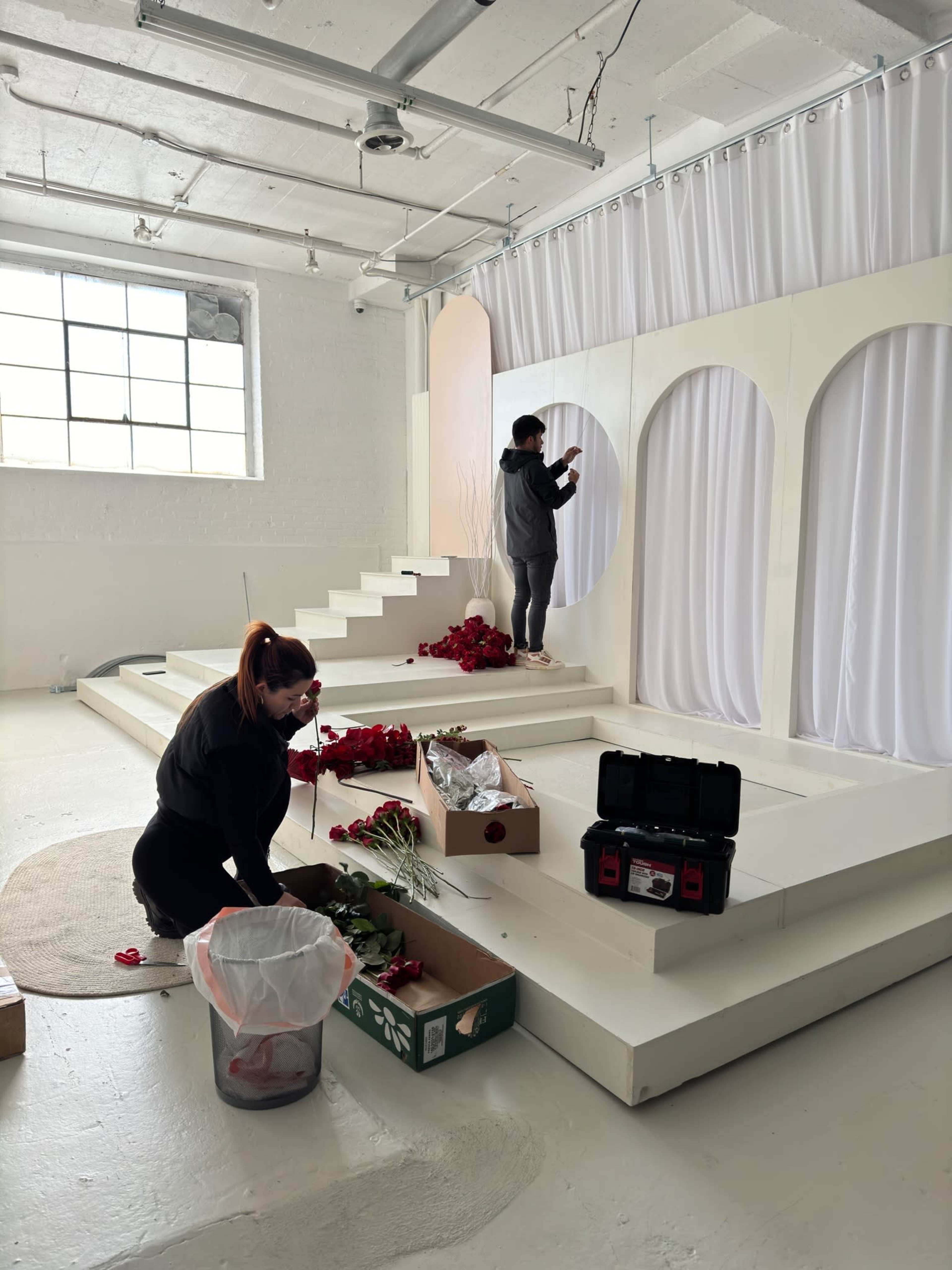 Two individuals are arranging flowers and adjusting drapery on a staged set with white steps and a large window.