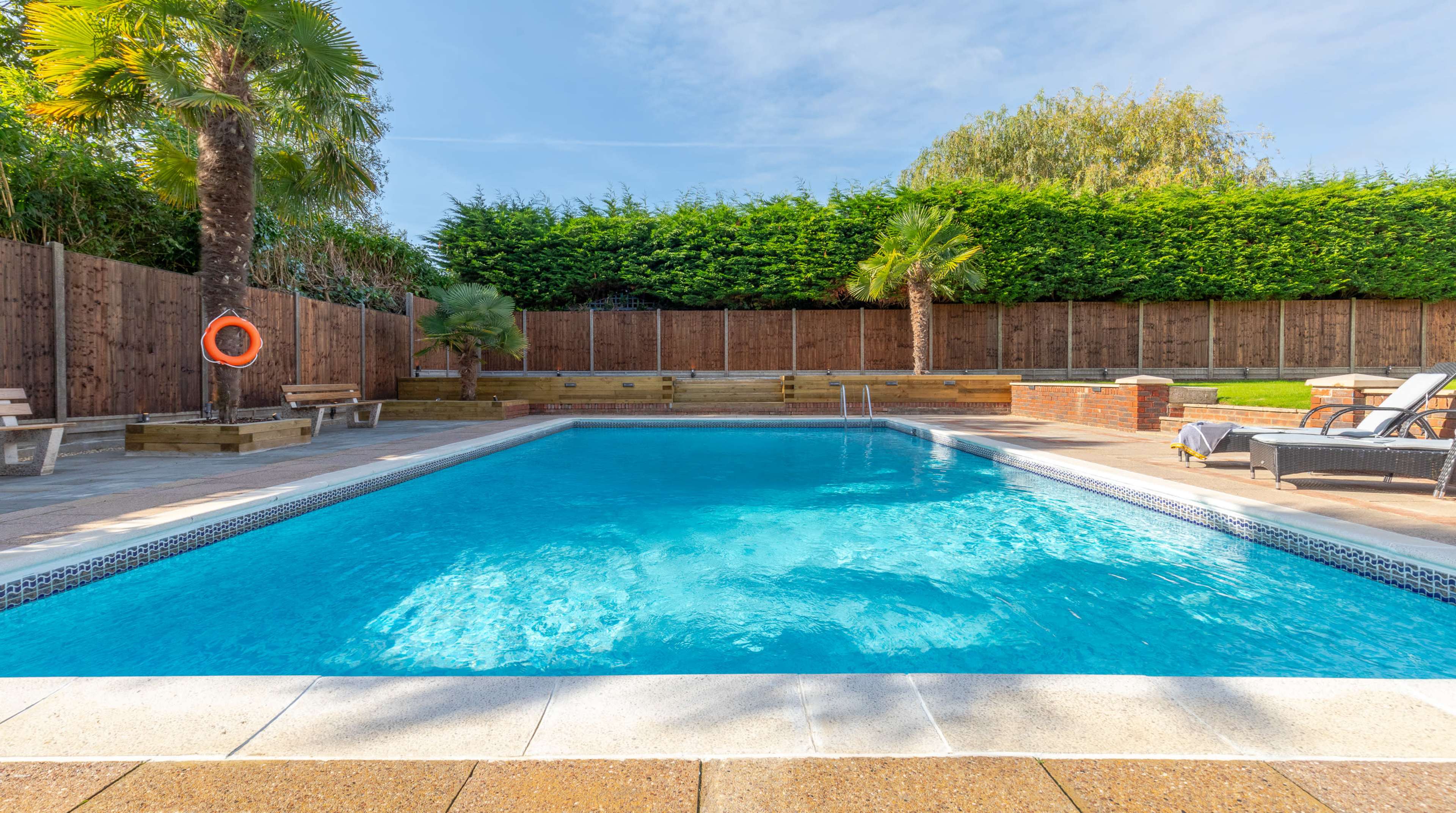 A clear swimming pool surrounded by stone decking, palm trees, and wooden fencing.