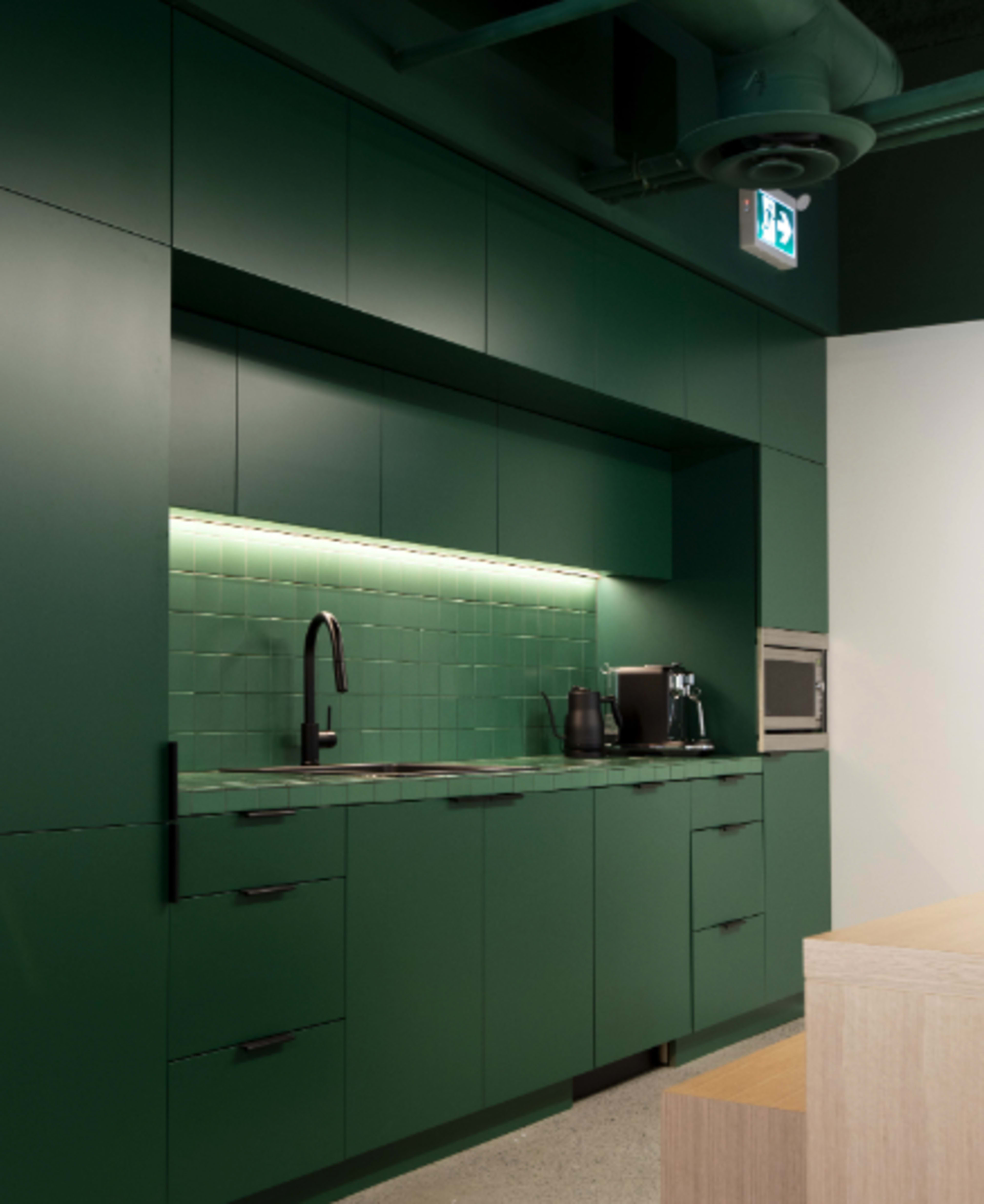 The image shows a modern kitchen with dark green cabinets, a backsplash of square tiles, and stainless steel appliances.