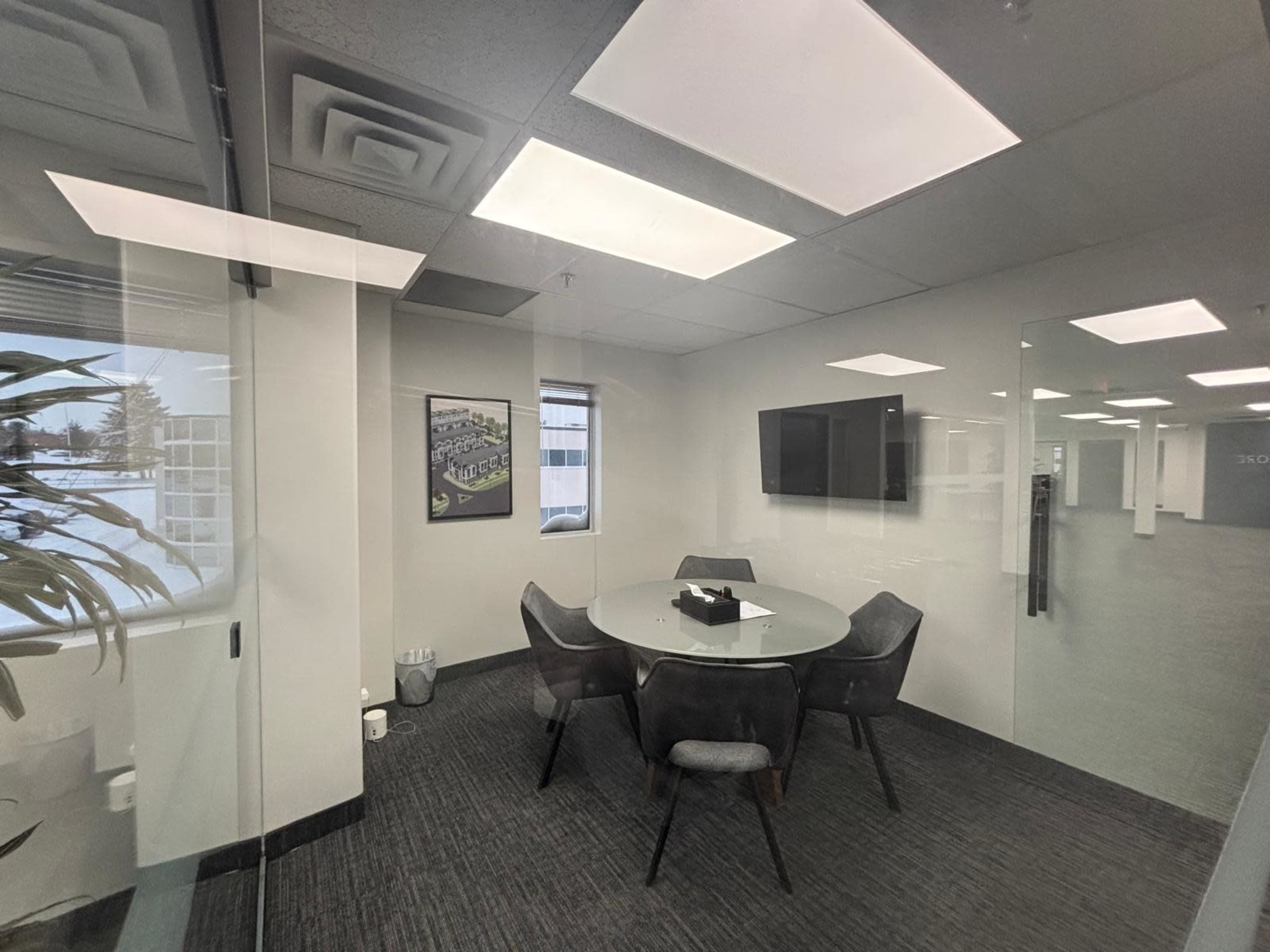 The image shows a modern conference room with a circular glass table surrounded by four chairs, a television on the wall, and large windows allowing natural light.