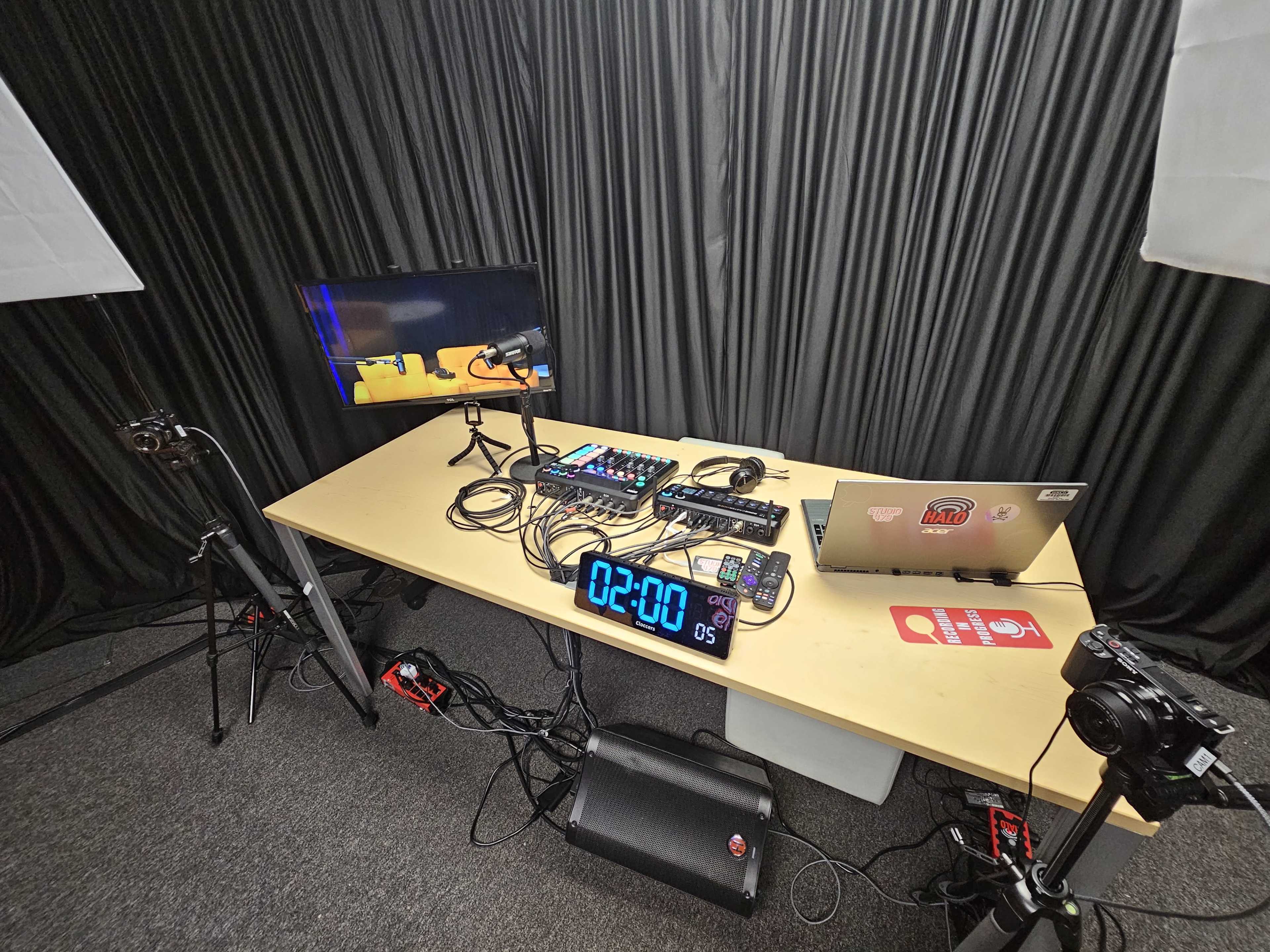 A tabletop setup for a recording studio features microphones, a laptop, audio equipment, and a timer display, with cameras positioned around the area.