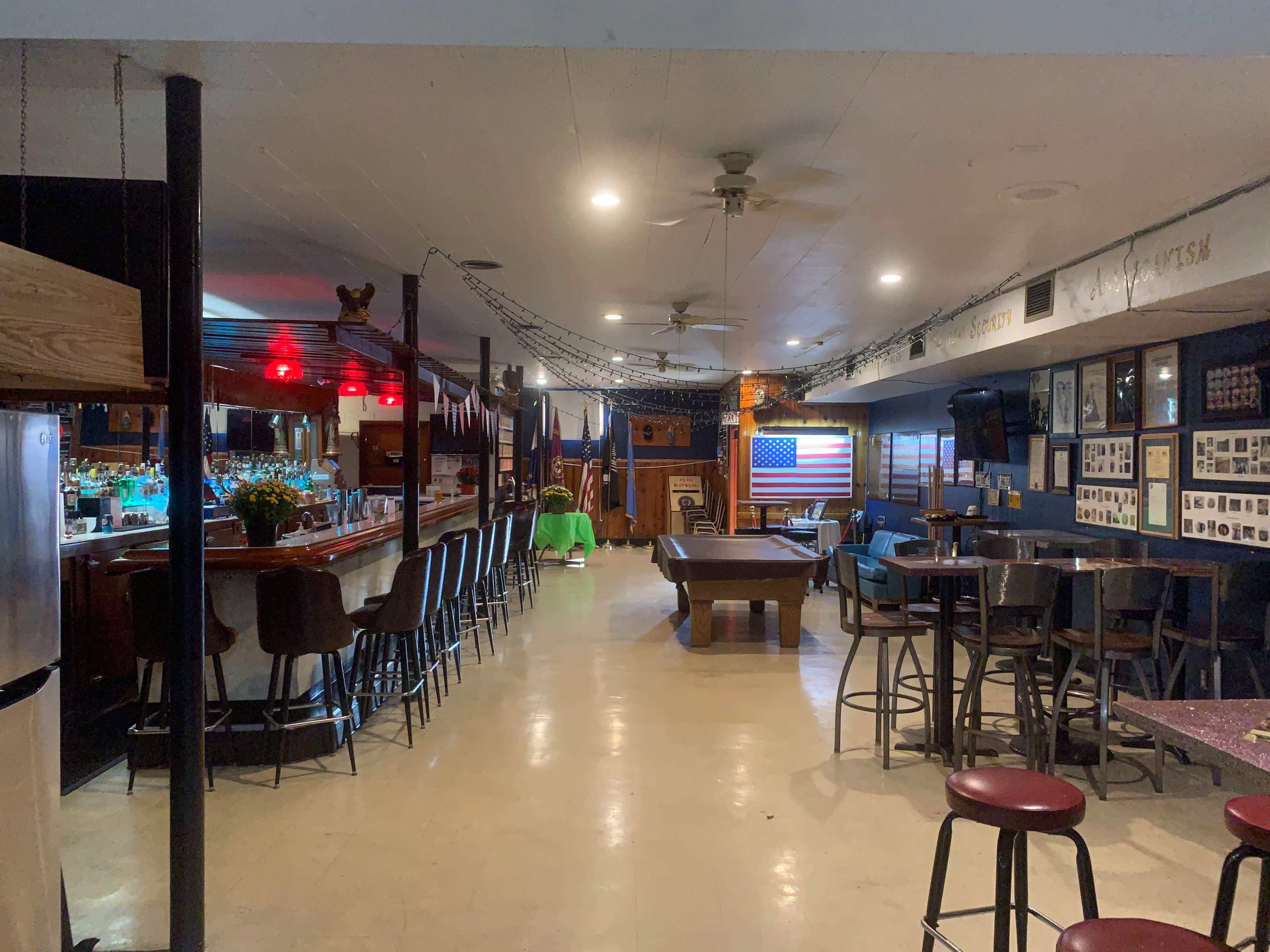 The interior of a bar features a pool table, a long counter with stools, and an American flag displayed on one wall.