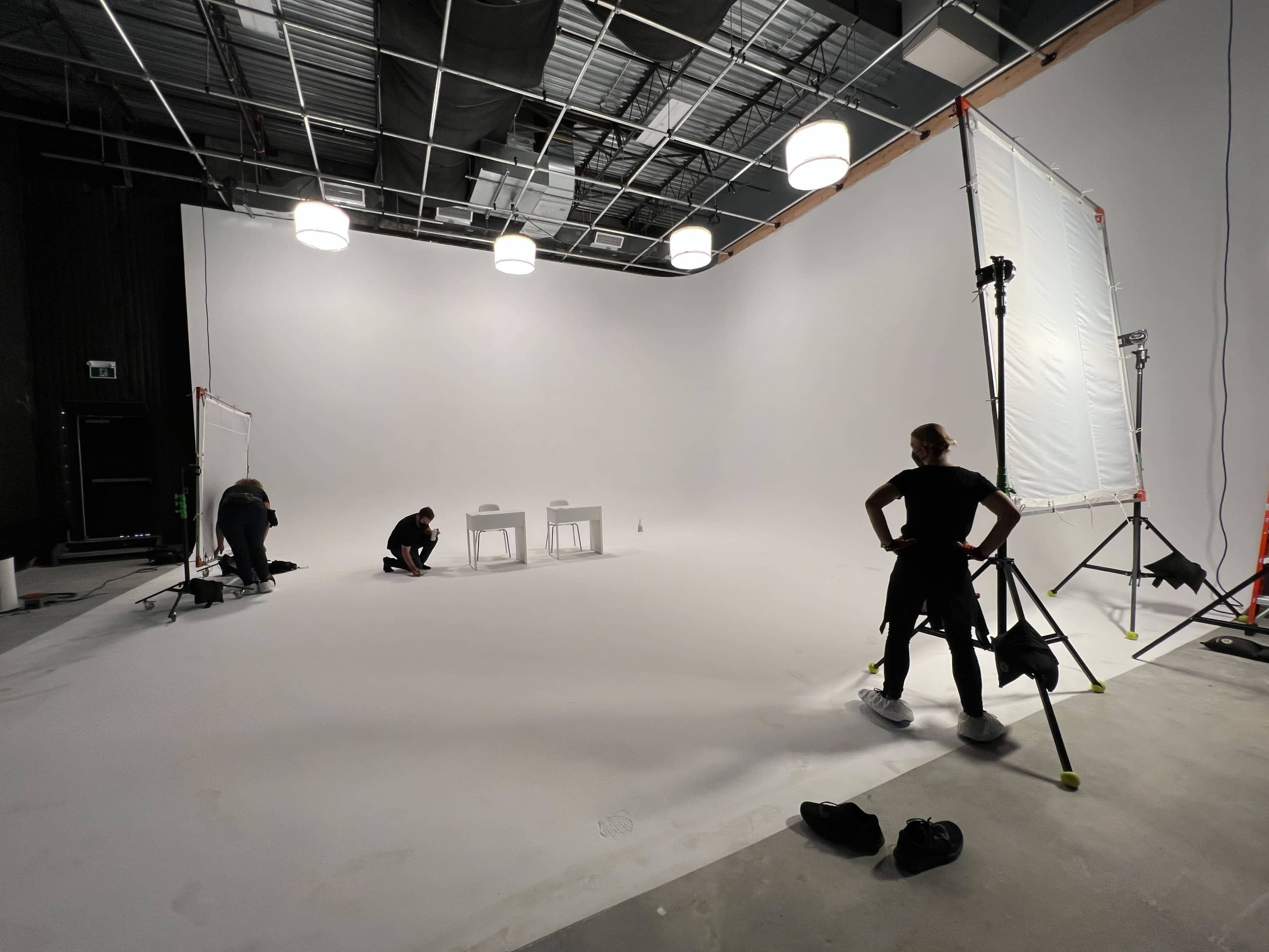 A large, empty studio space is being prepared for a shoot, with two tables and several crew members working in the scene.