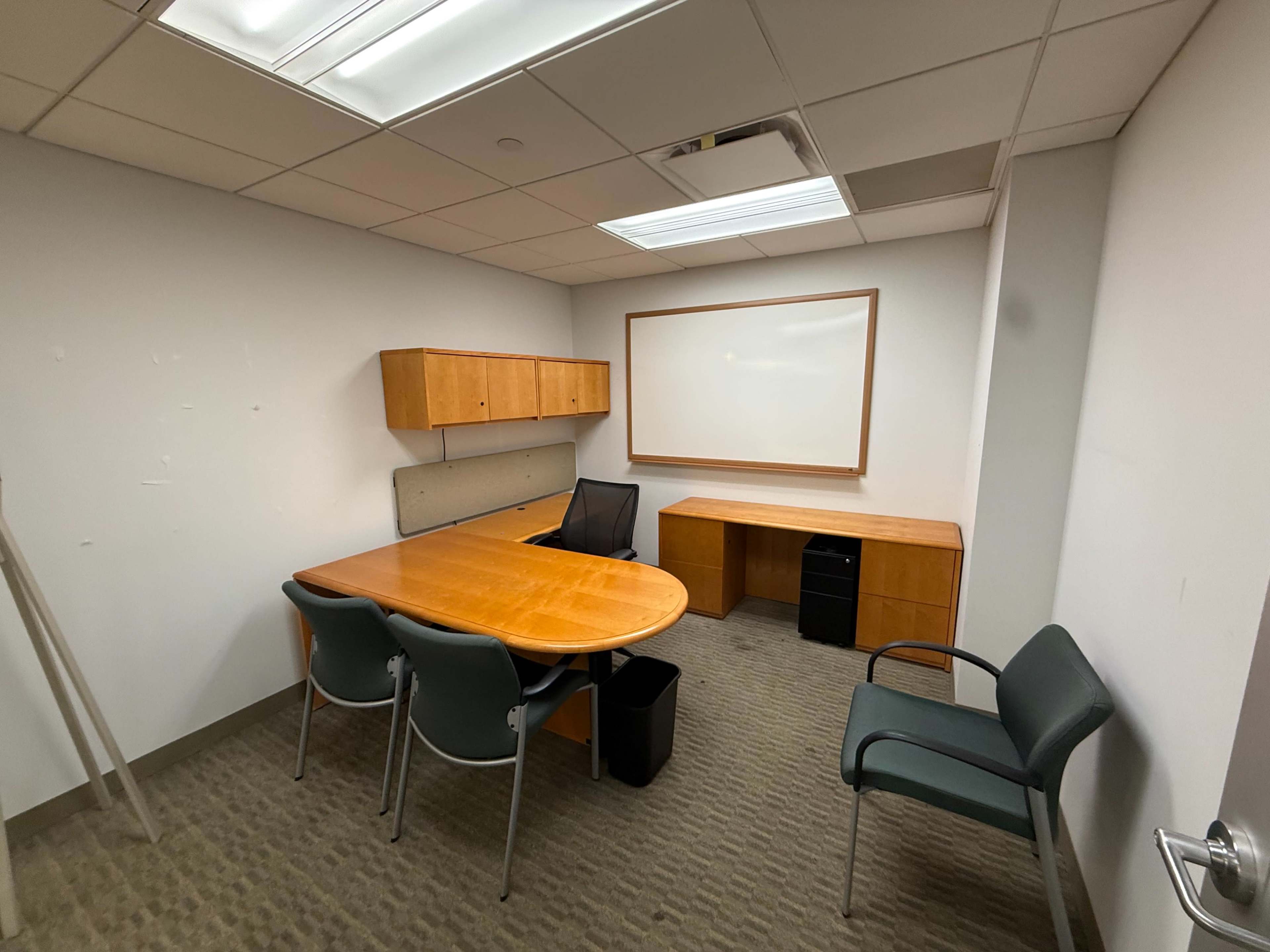 The image shows a simple, empty office with a wooden table, black chair, and wall-mounted whiteboard.