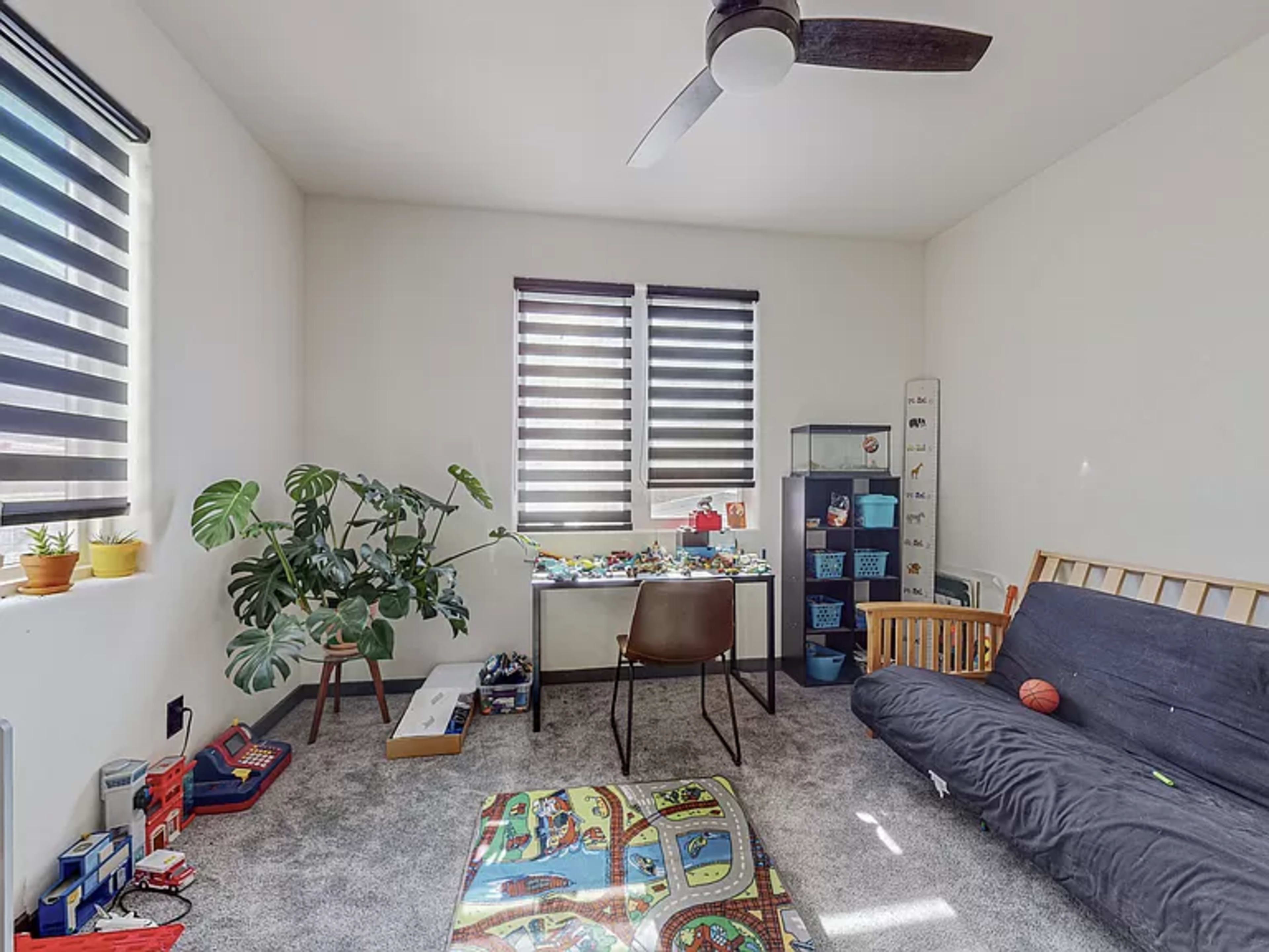 A children's room with a sofa bed, a table surrounded by toys, a large plant, and storage bins against the wall.