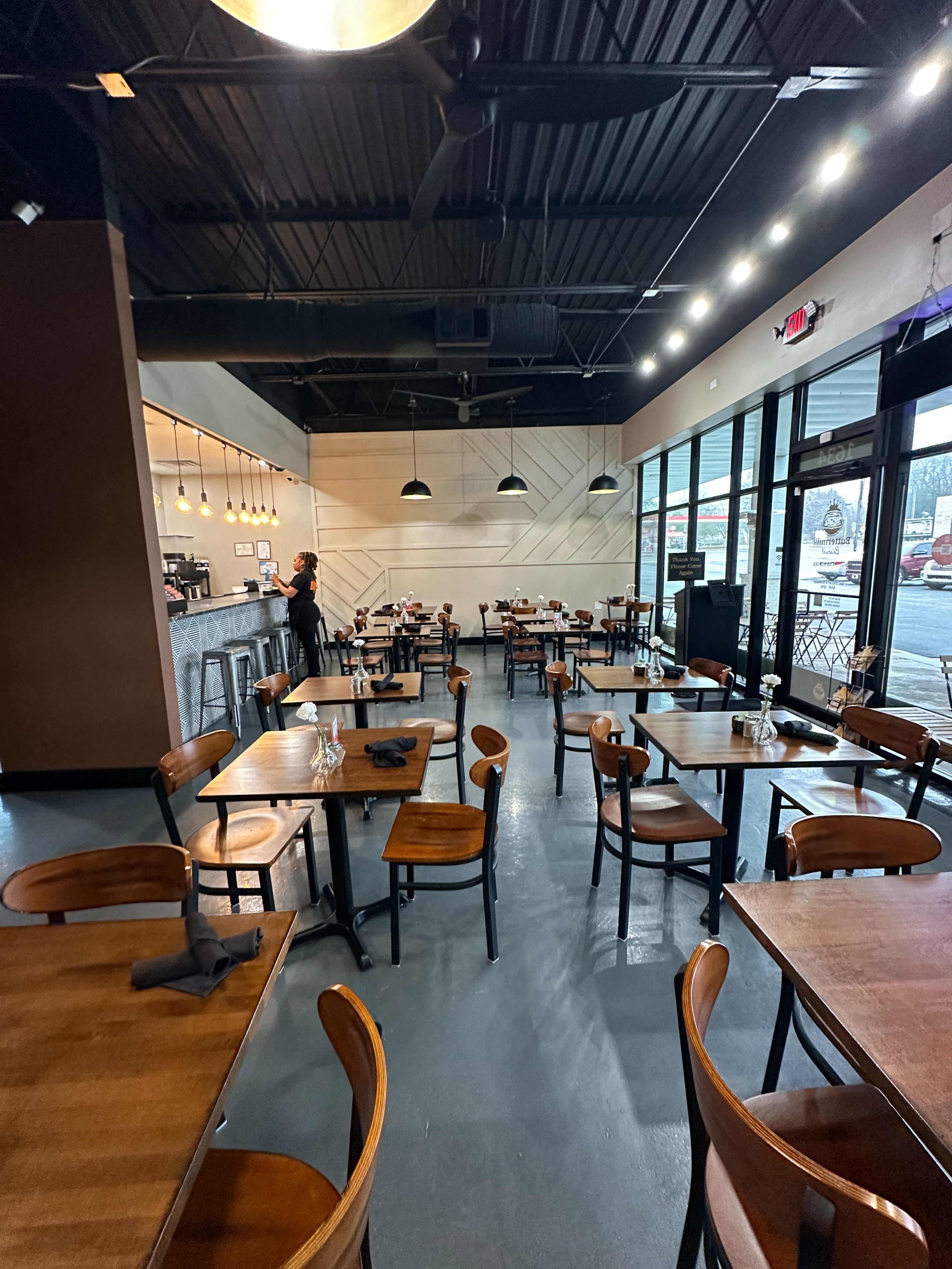 A spacious restaurant interior with wooden tables and chairs arranged neatly, and a staff member standing near the counter.