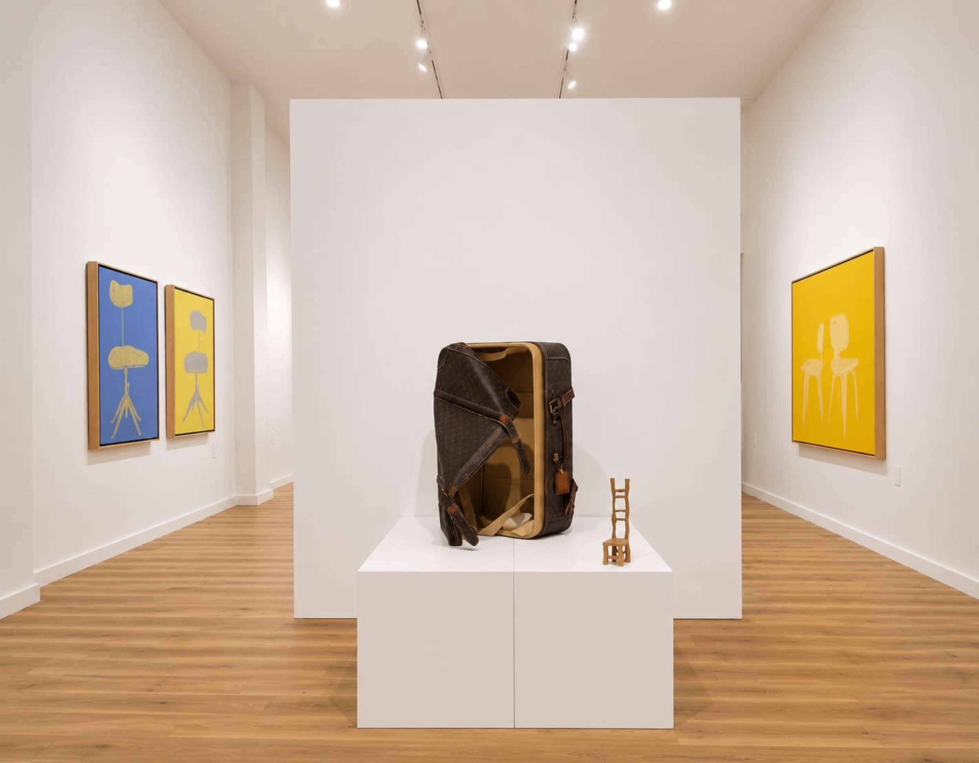 The image shows an art gallery featuring a wooden display pedestal with a brown backpack and a small wooden sculpture, surrounded by framed paintings on the walls.
