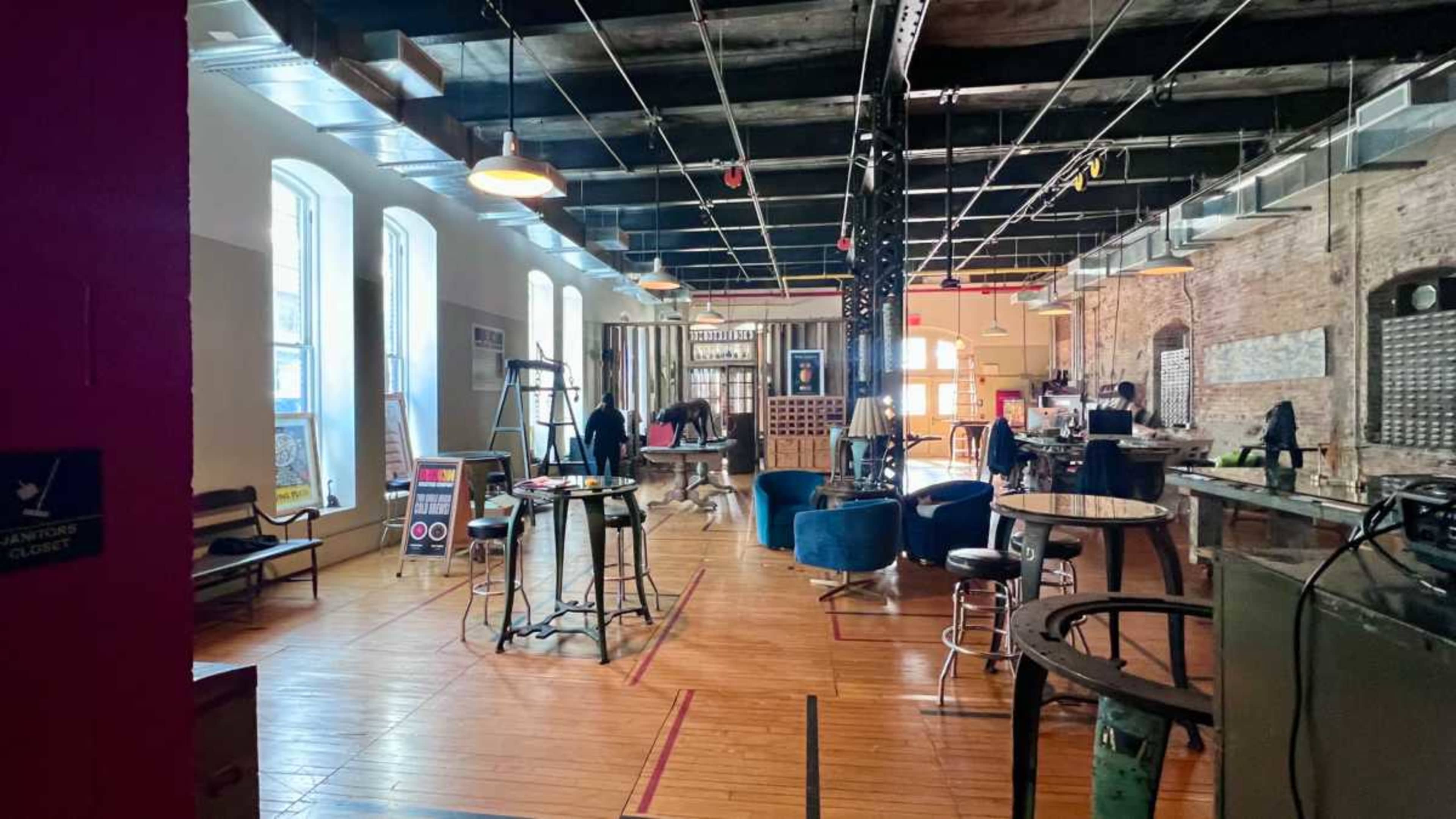 The image shows an open, spacious interior of a café or community space with various seating arrangements, exposed brick walls, and large windows allowing natural light.