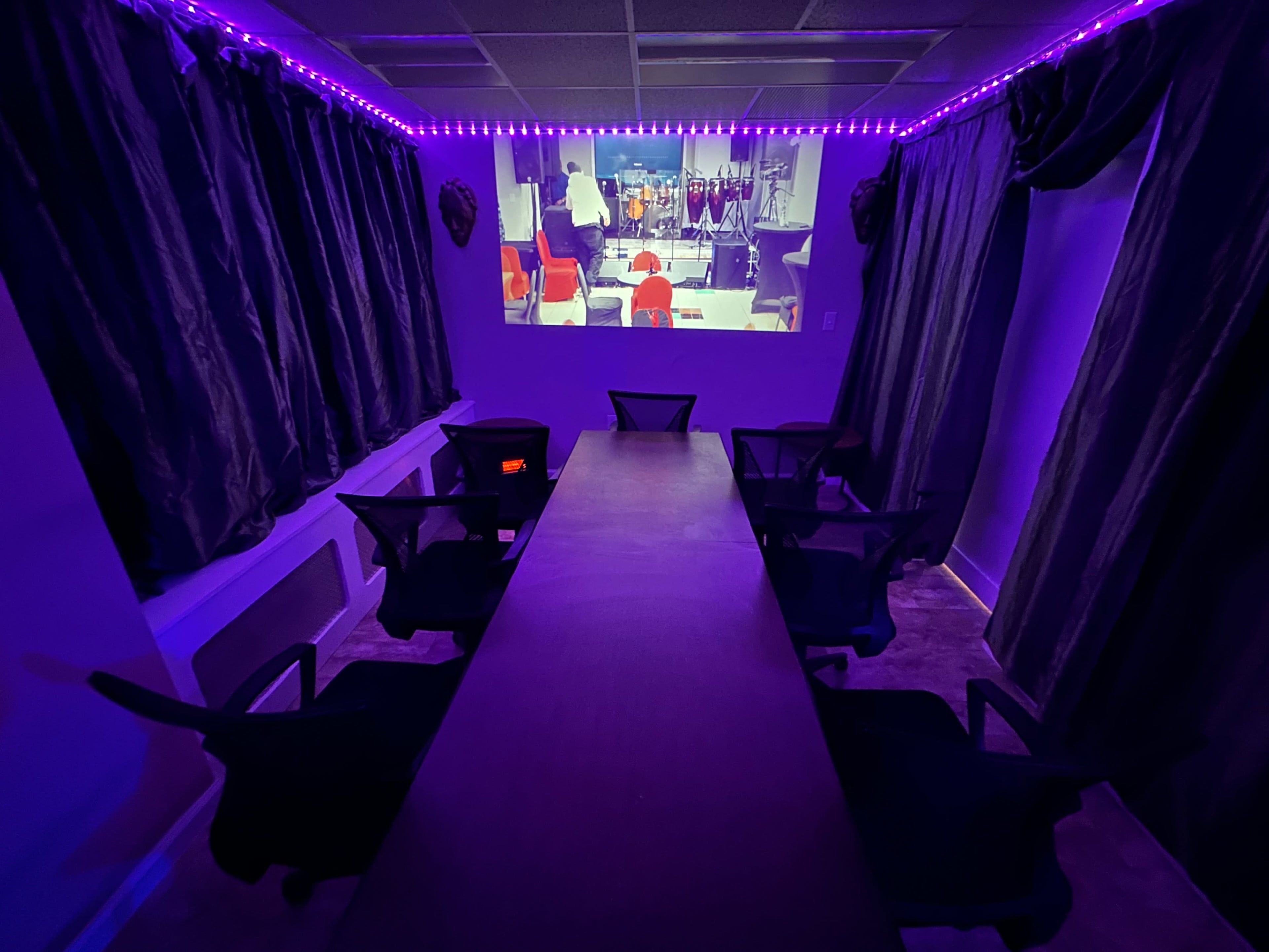 A dimly lit meeting room features a long table surrounded by chairs, with a large projection screen displaying a video in the background and purple LED lights illuminating the space.