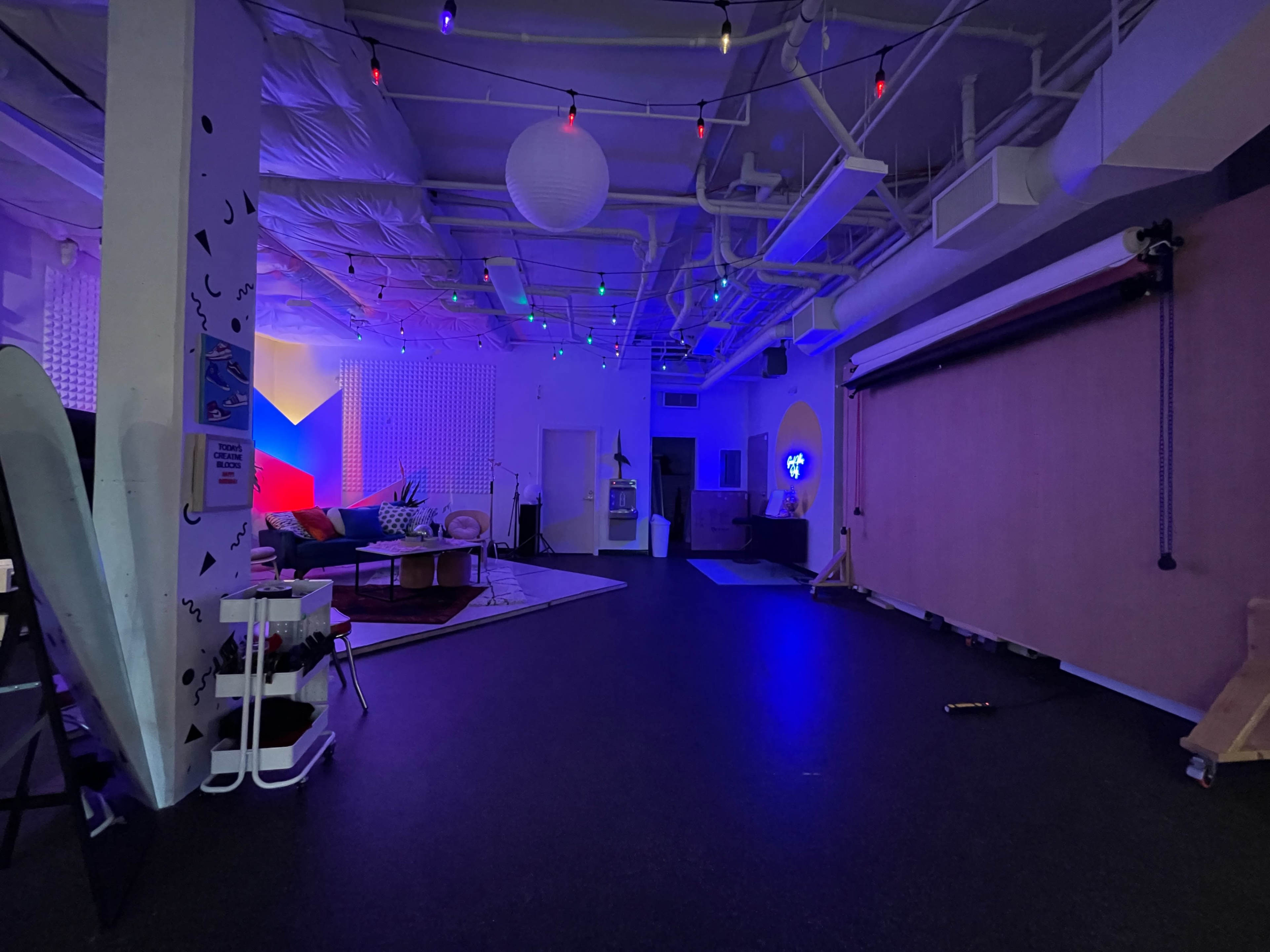 The image shows a dimly lit room with colorful lights, a large wall-mounted backdrop, and a seating area featuring a couch and decorative items.