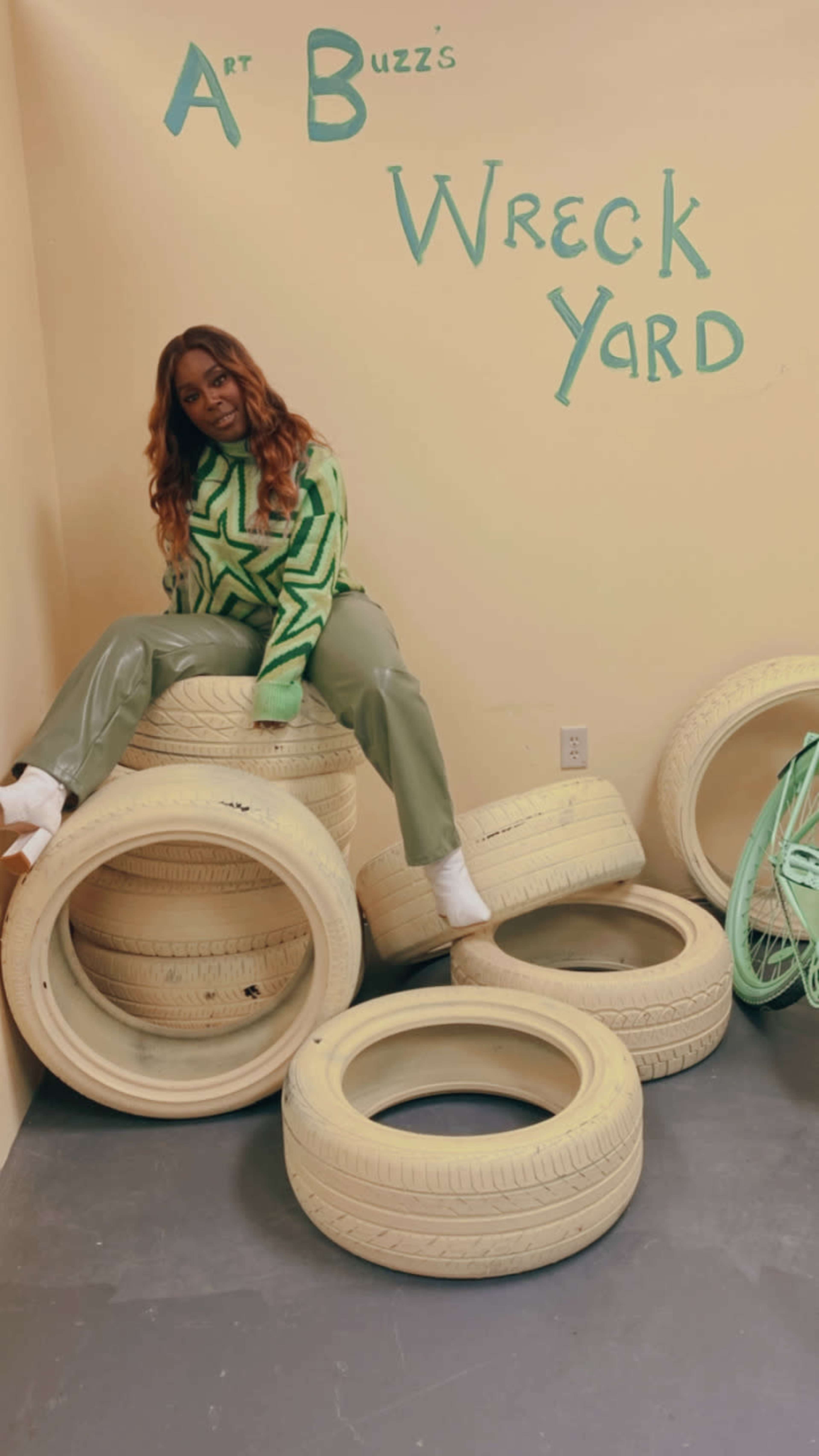 A person wearing a green star-patterned sweater and glossy pants sits atop a stack of white tires in a room with "Art Buzz's Wreck Yard" painted on the wall.