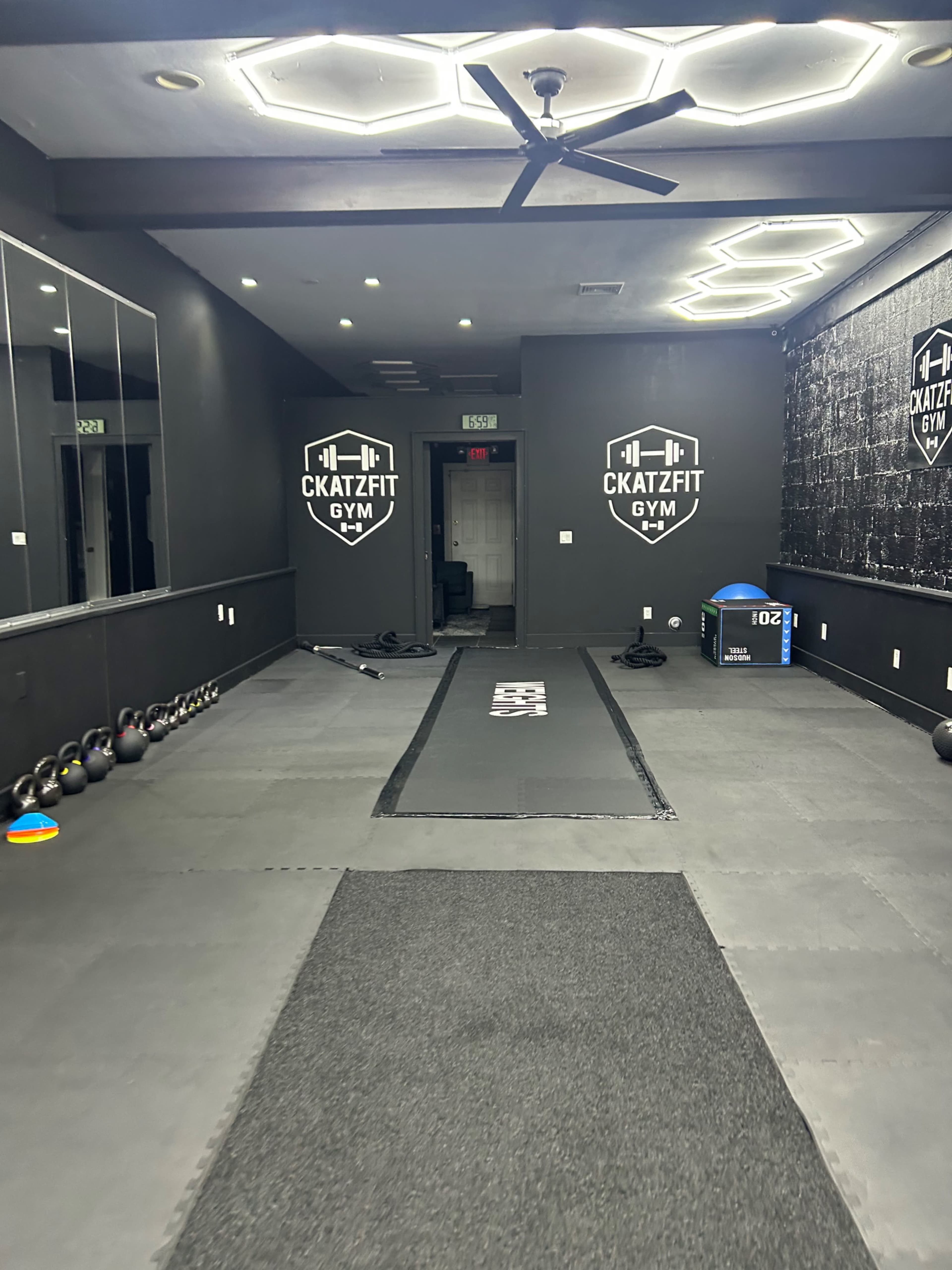The image shows the interior of a gym with a rubber floor, a central workout area, and wall-mounted logos for "KatzFit Gym."
