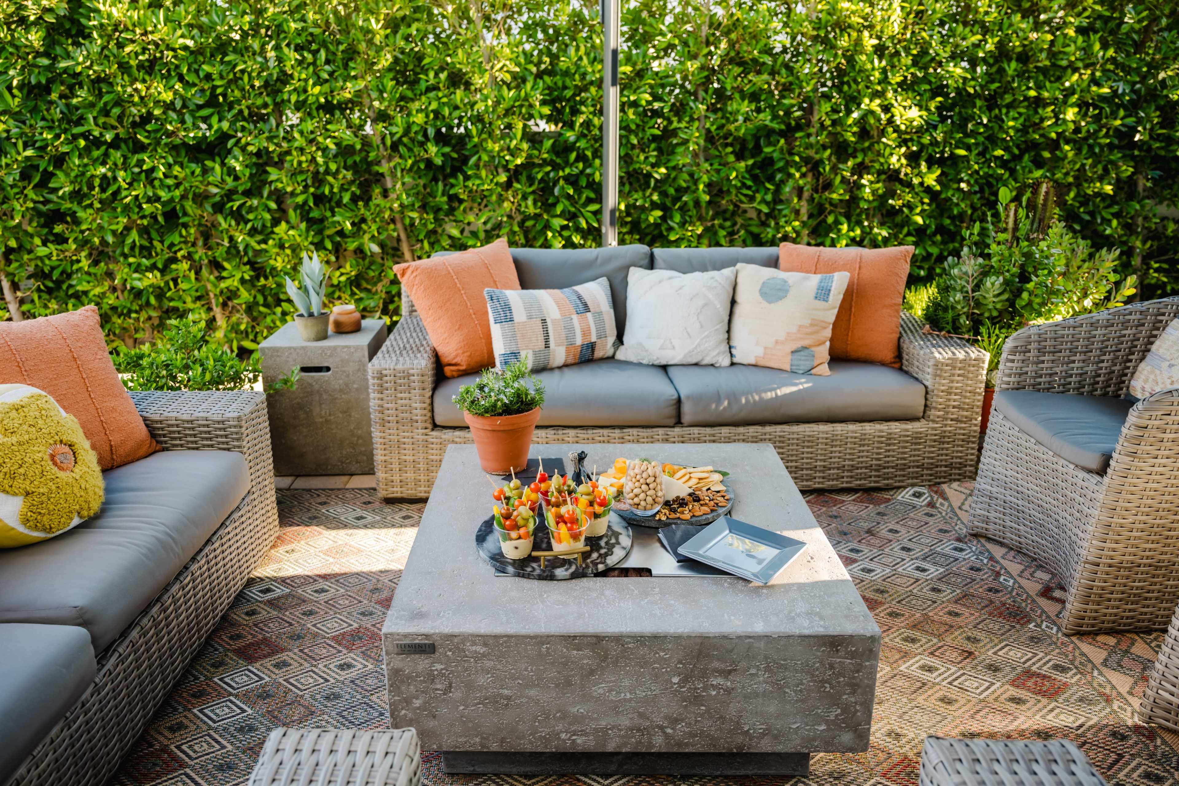 A cozy outdoor seating area features wicker furniture, decorative cushions, and a stone coffee table adorned with an assortment of snacks and drinks.