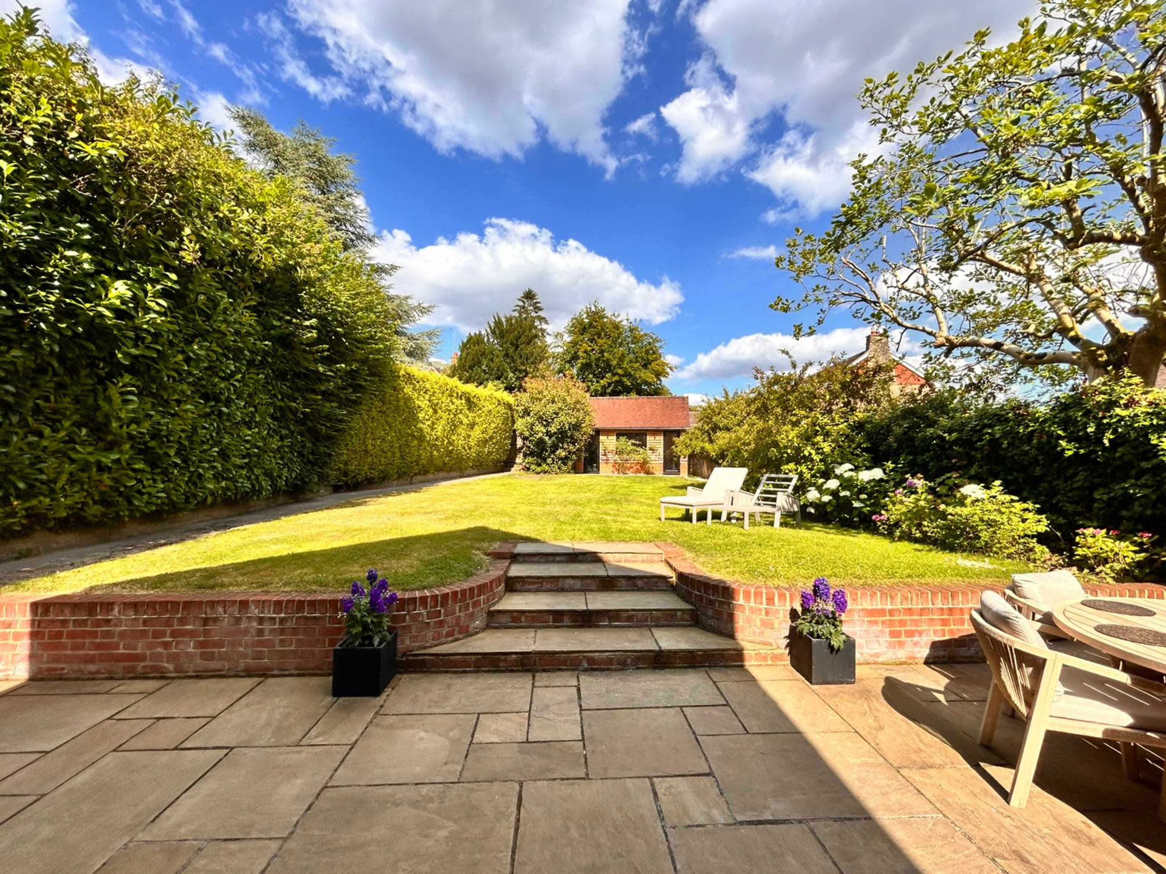 A well-maintained garden features lush green grass, a stone patio with steps, and a gazebo in the background under a partly cloudy sky.