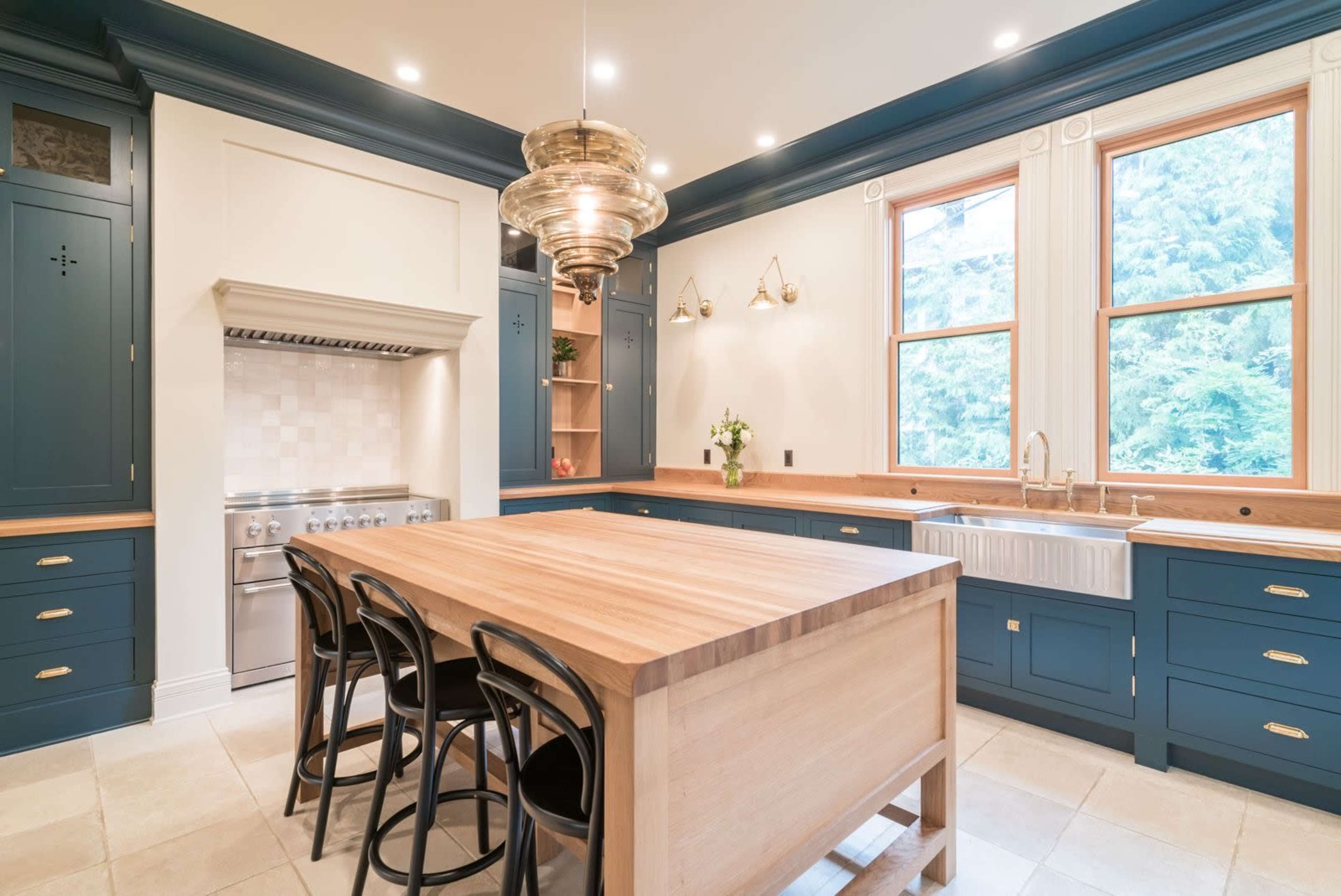The kitchen features blue cabinetry, a central wooden island with black chairs, and large windows that provide natural light.