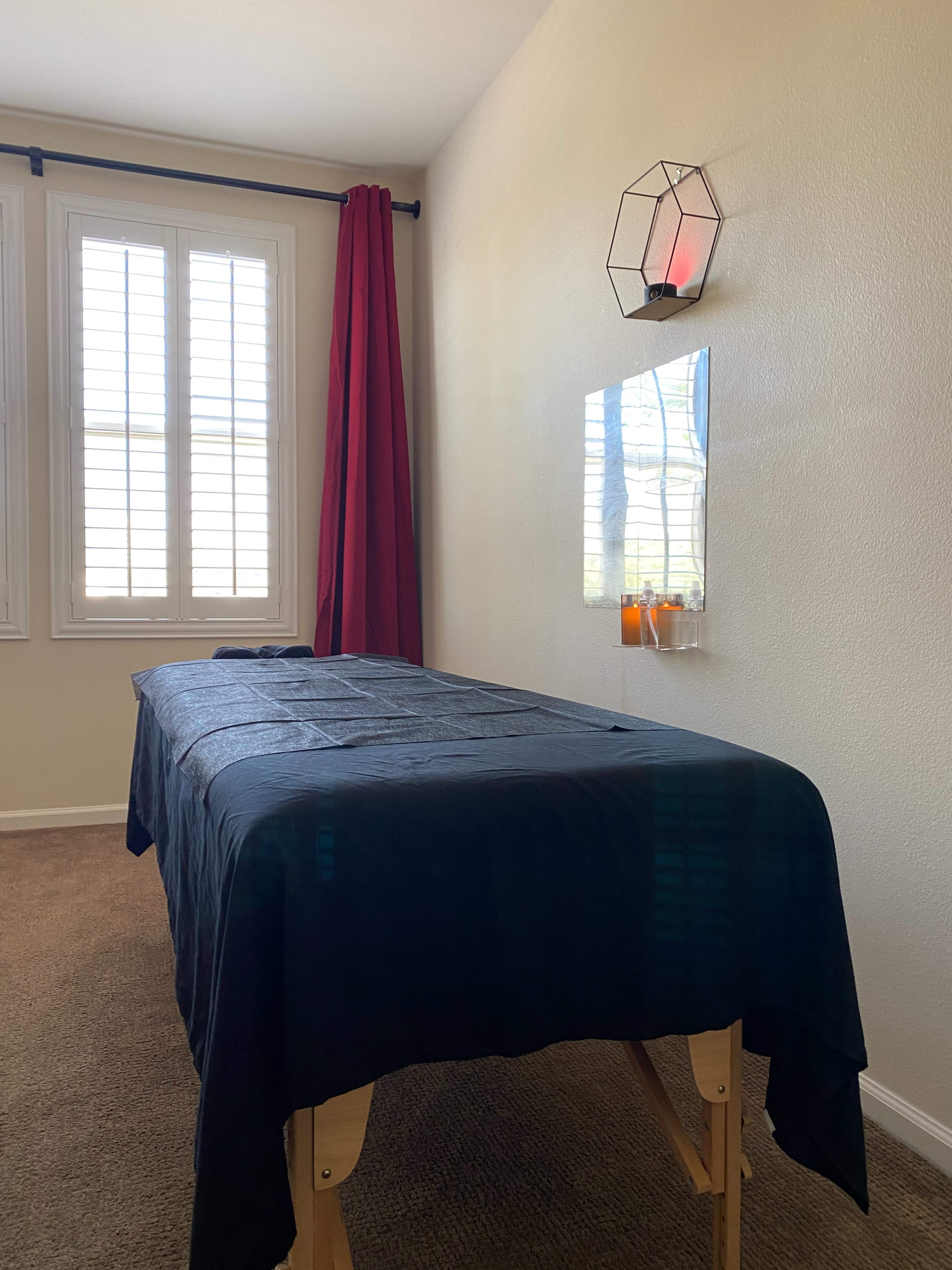 A massage therapy room with a massage table covered in a dark cloth, a red curtain, and a wall-mounted candle holder beside a mirror.