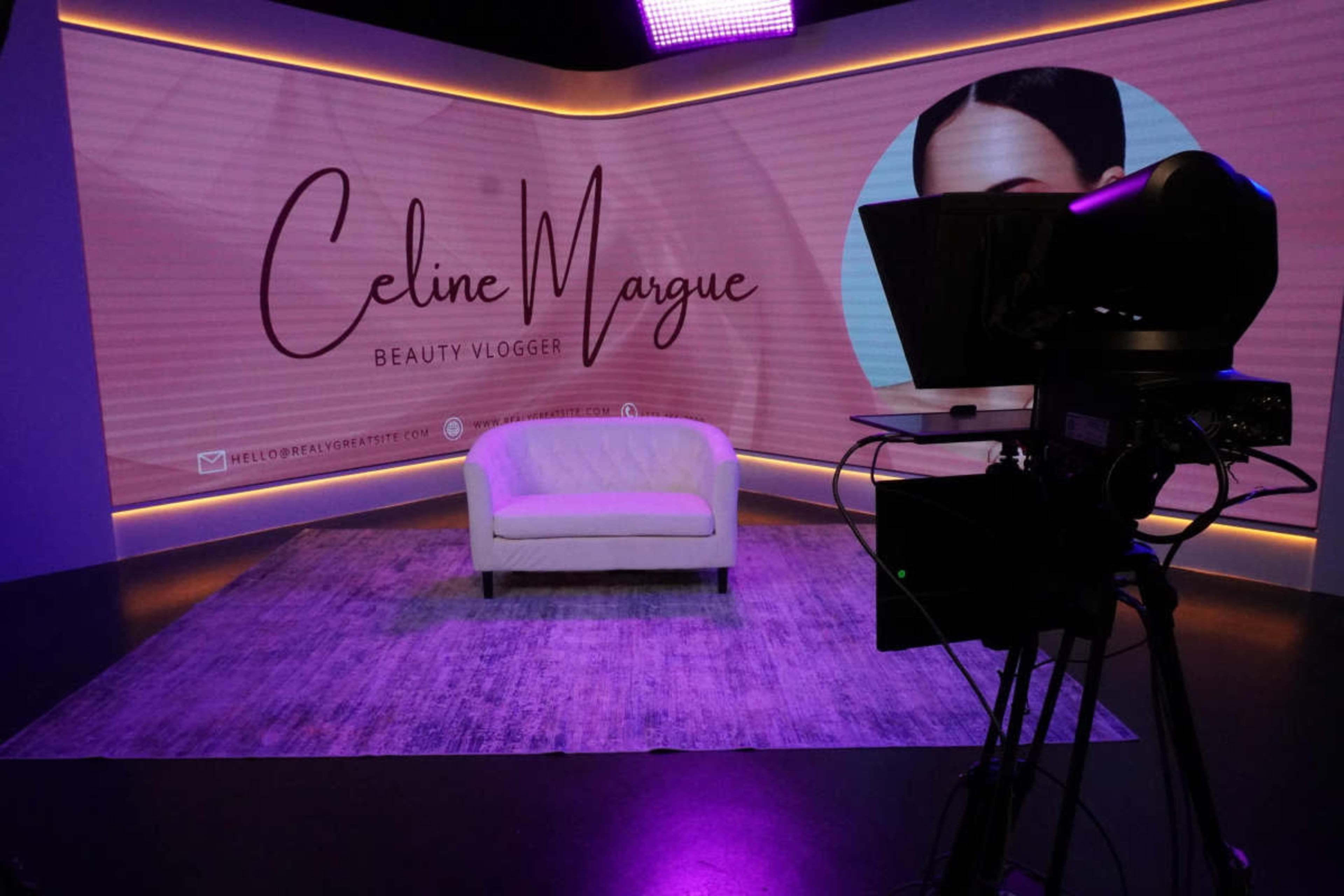 A white sofa is positioned on a patterned rug in a studio set featuring the name "Celine Margue" and the title "Beauty Vlogger" on a large backdrop.