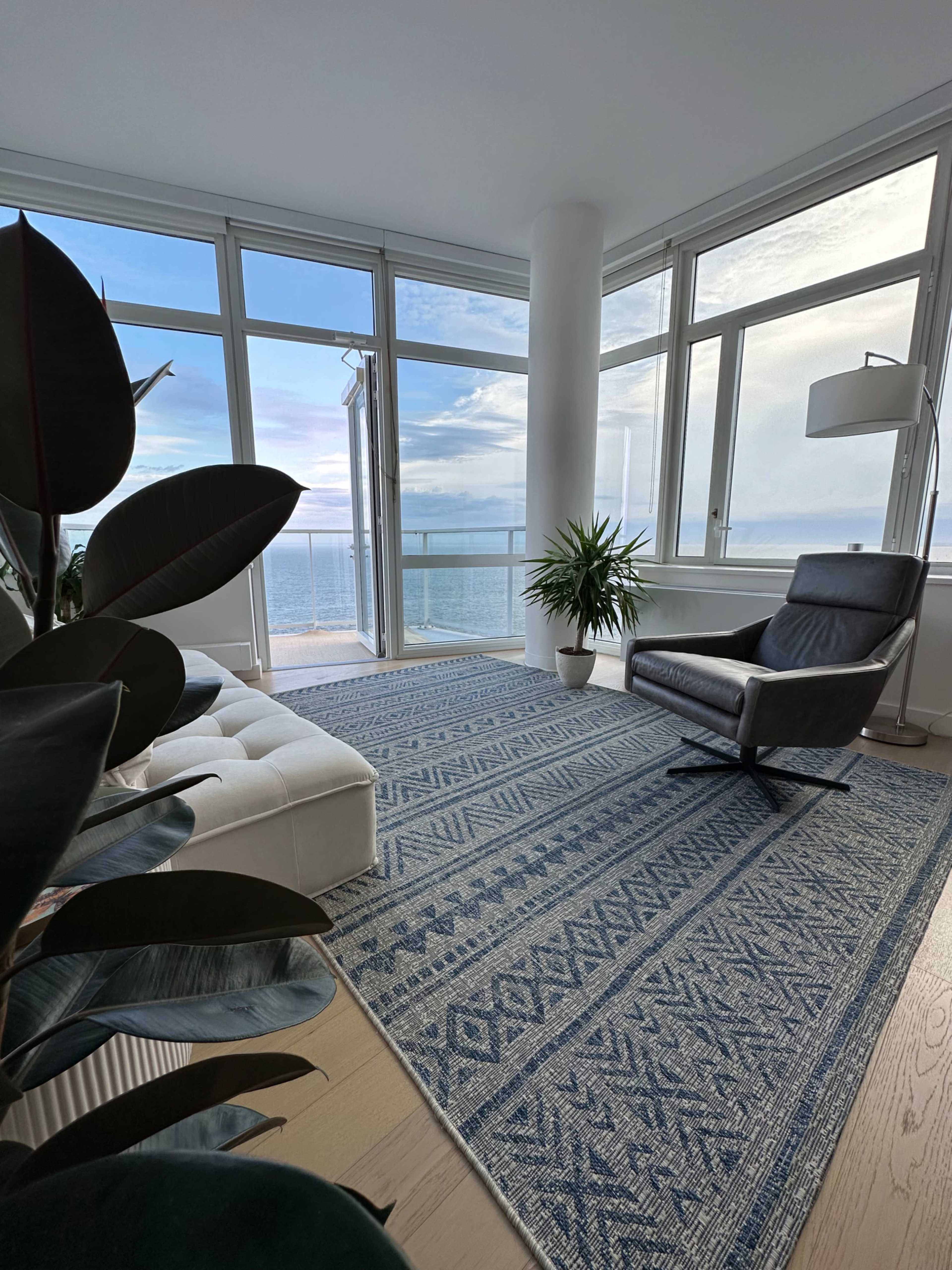 A modern living room features a large window with a view of the ocean, a black armchair, a potted plant, and a patterned blue rug on wooden flooring.