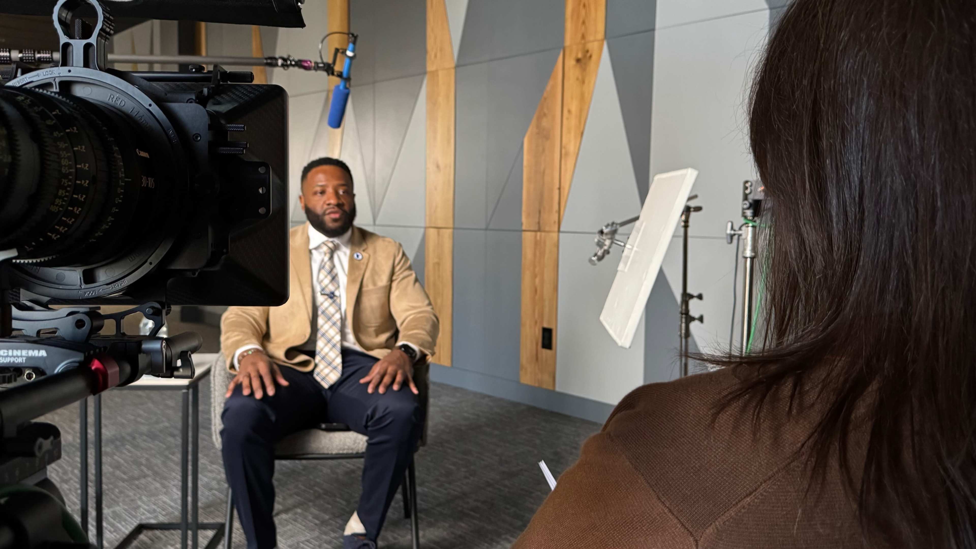 A man in a tan blazer sits in front of a camera, speaking during an interview in a well-lit room with wooden accents.