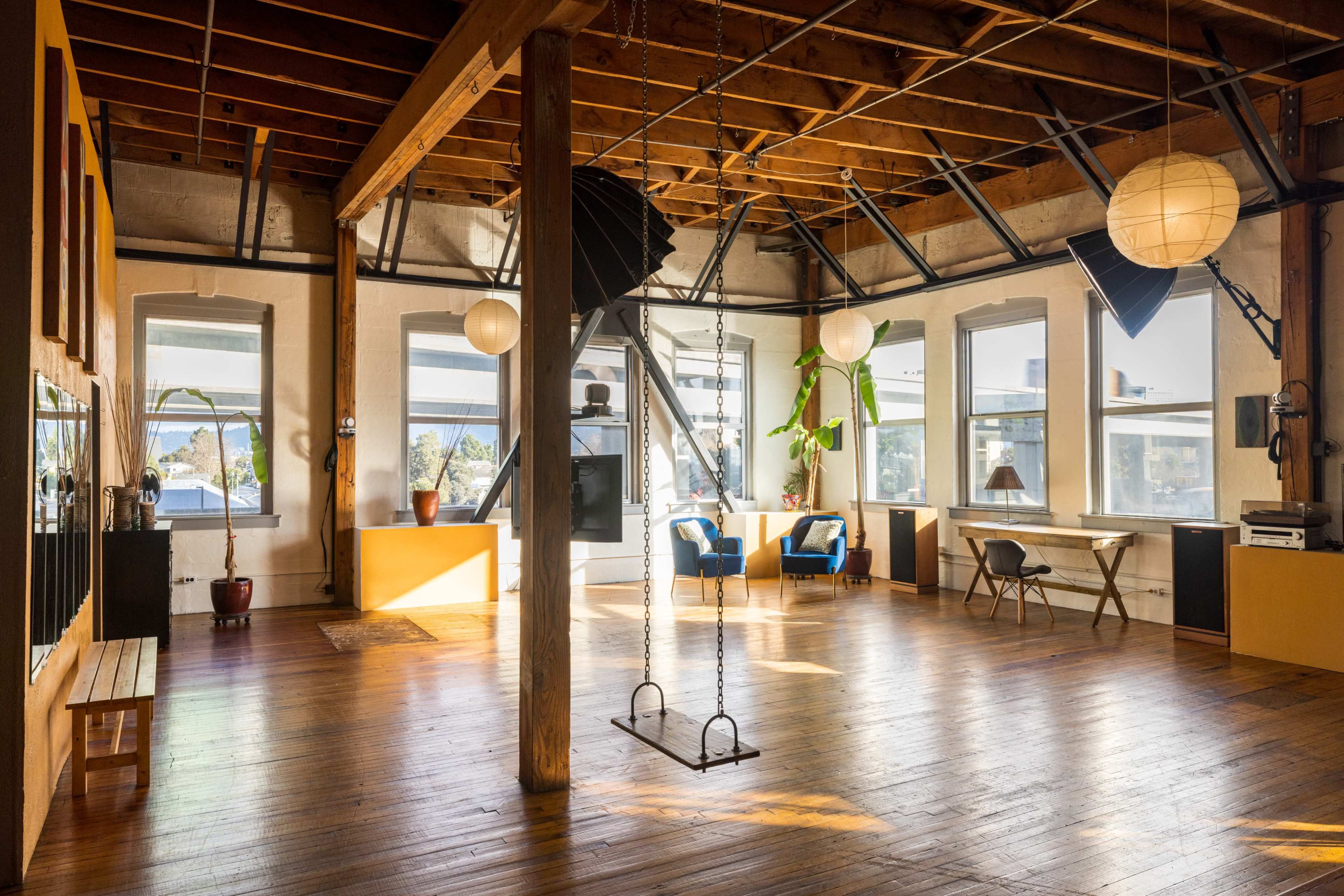 The image shows a spacious loft with high wooden ceilings, large windows, and a swing hanging from a beam, alongside various furniture pieces and plants.