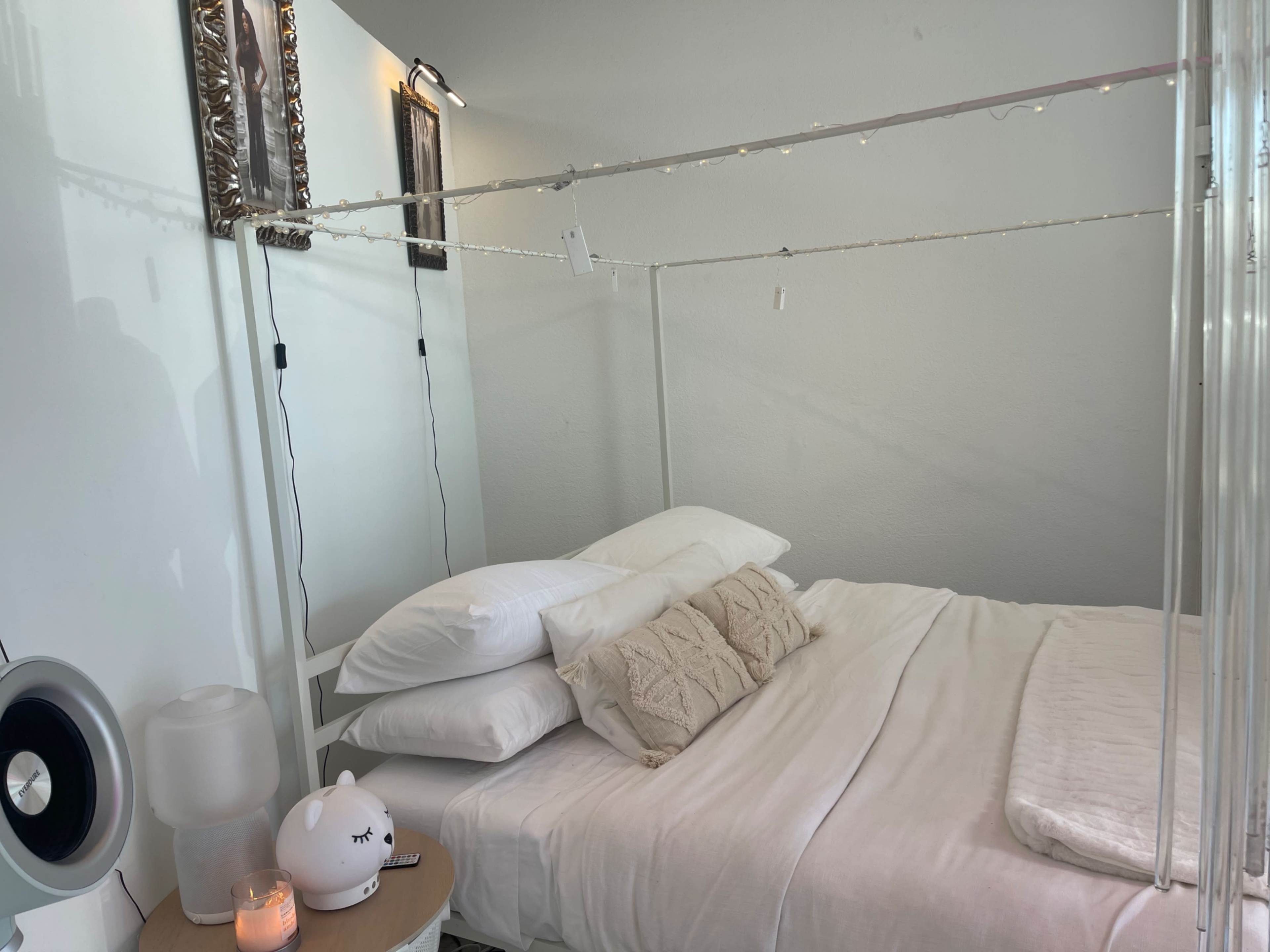 A bed with a canopy frame is positioned in a softly lit room, featuring white bedding and decorative pillows.