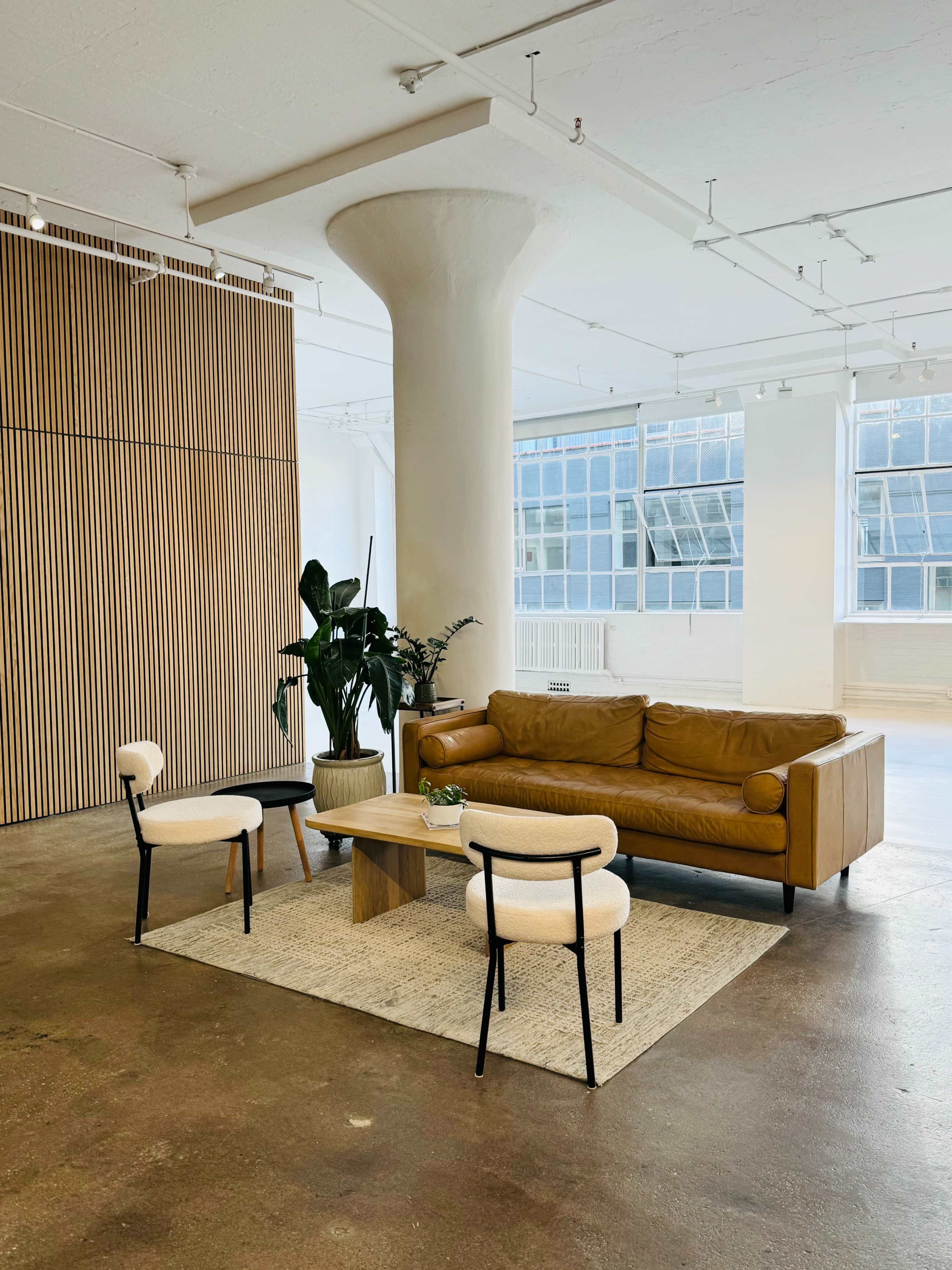 The image shows a spacious, modern living area featuring a brown leather sofa, two white chairs, a wooden coffee table, and a large indoor plant, all set against a backdrop of large windows and wooden panels.