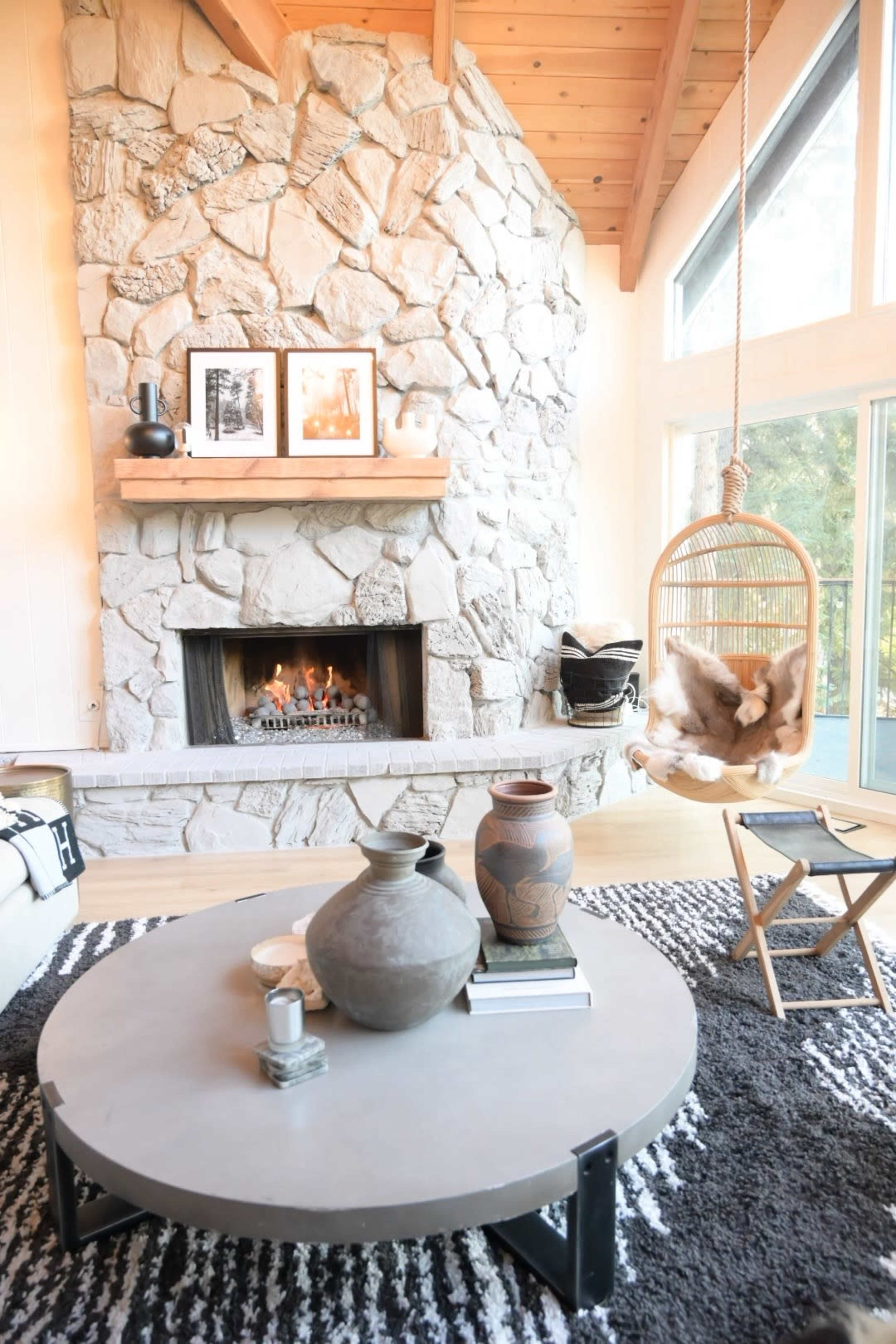 A cozy living room features a stone fireplace with a mantel displaying framed photos, a round coffee table, and a hanging chair beside a large window.