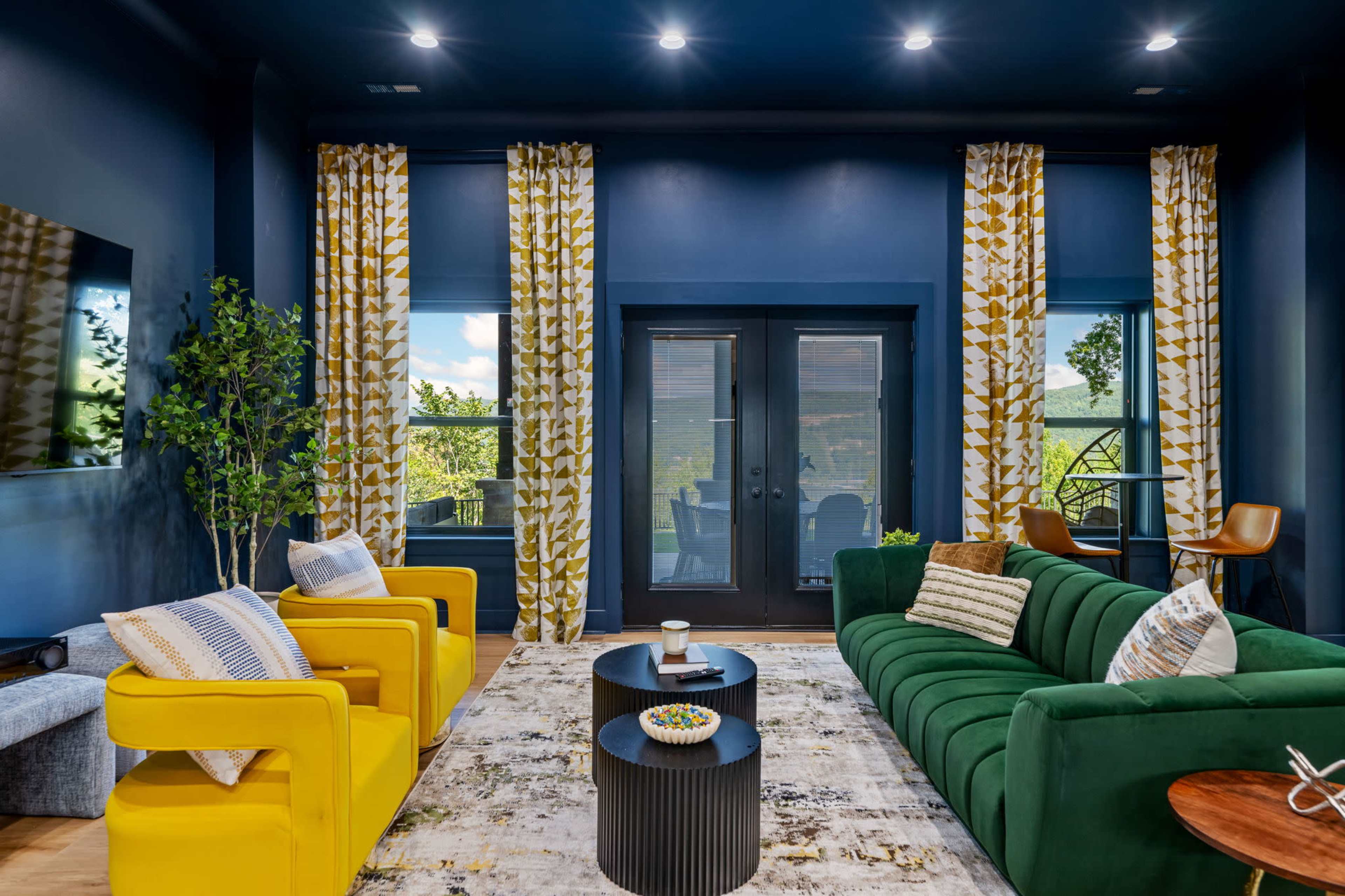 The image shows a modern living room featuring a green sofa, a yellow armchair, patterned curtains, and a coffee table with decorative items.