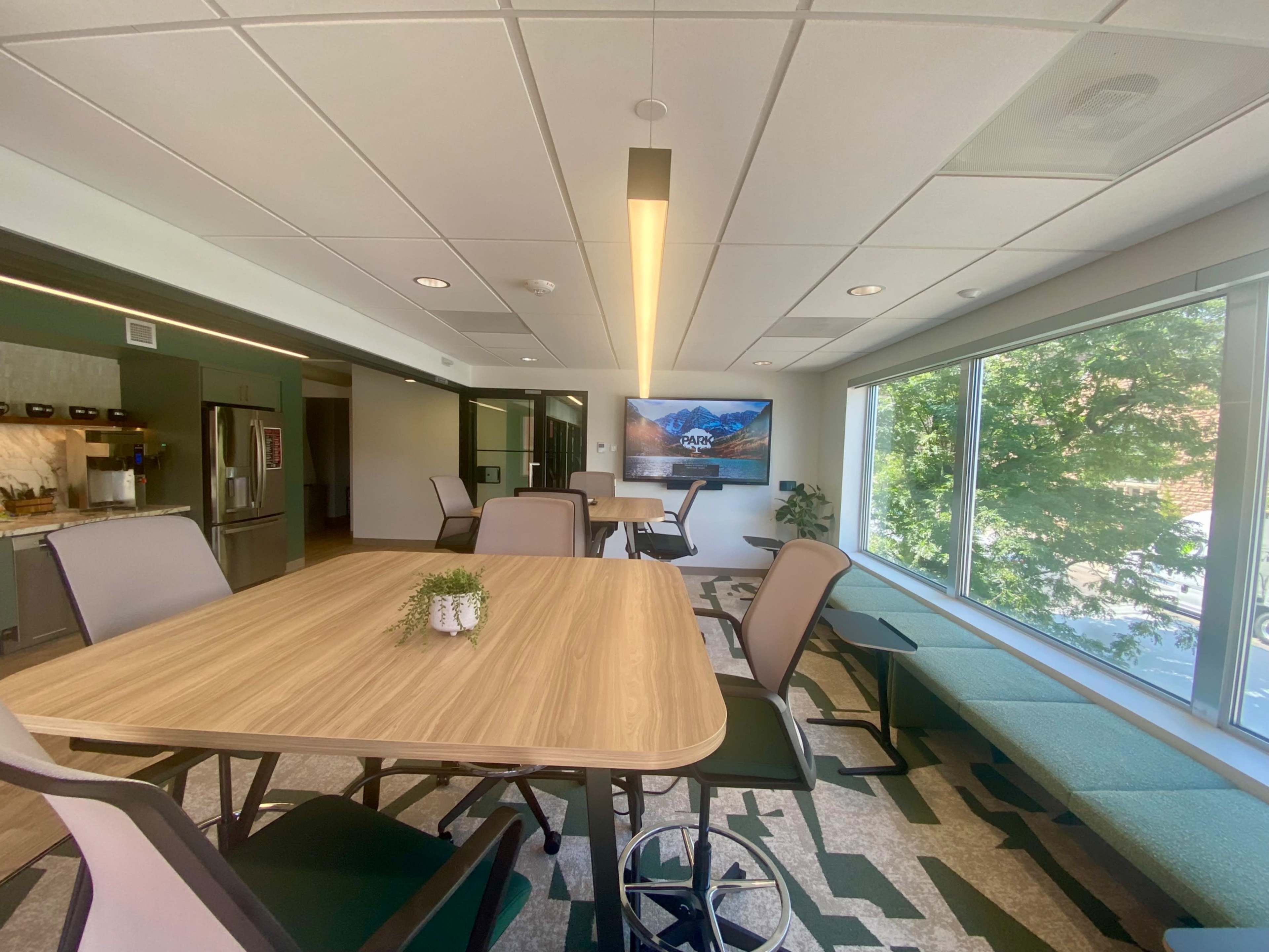 The image shows a modern office space with wooden tables, ergonomic chairs, and large windows allowing natural light to fill the room.