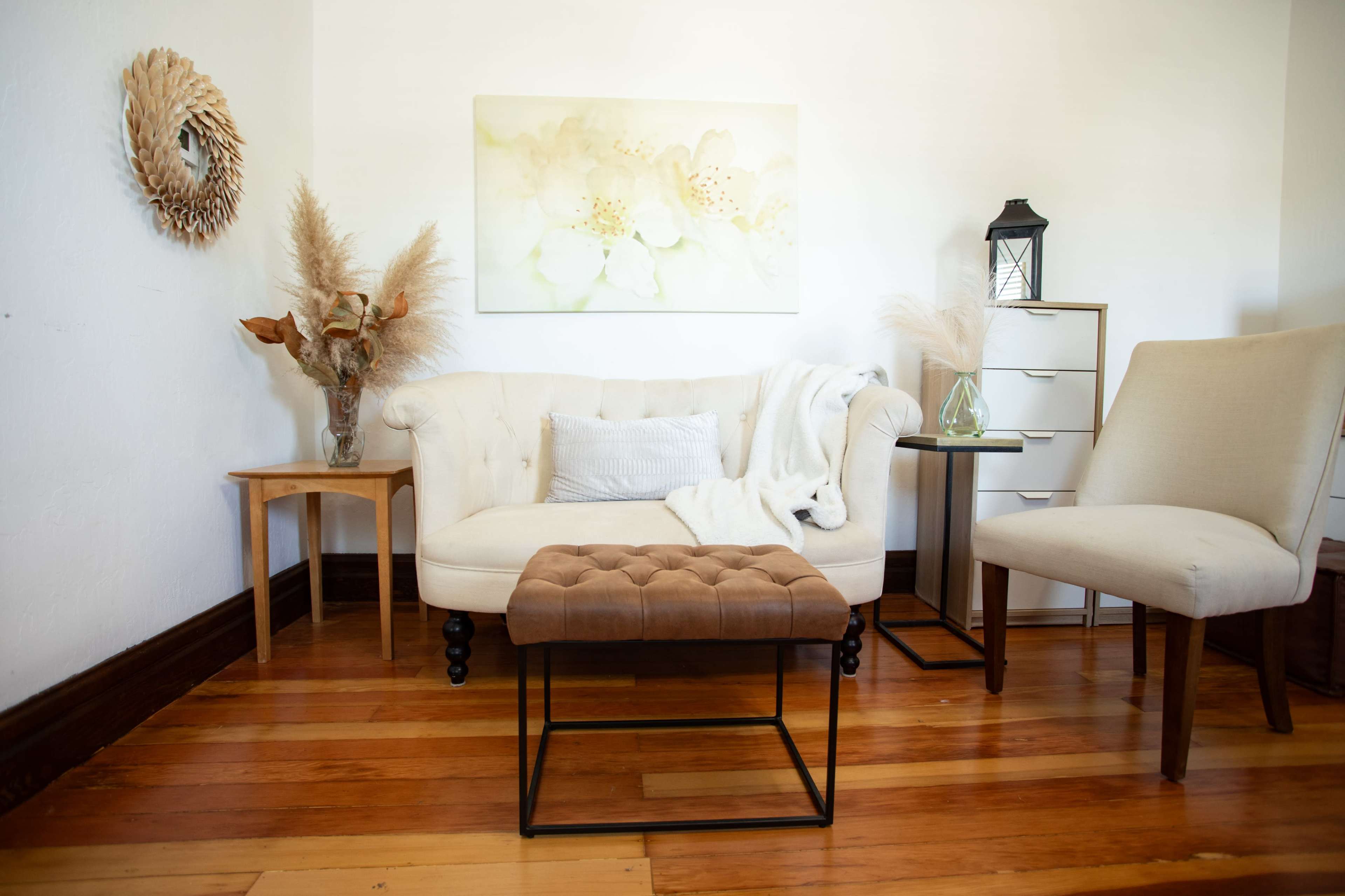 A light and airy living room features a beige tufted ottoman, a white sofa with a throw blanket, and decor elements such as a vase with dried flowers and a small side table.