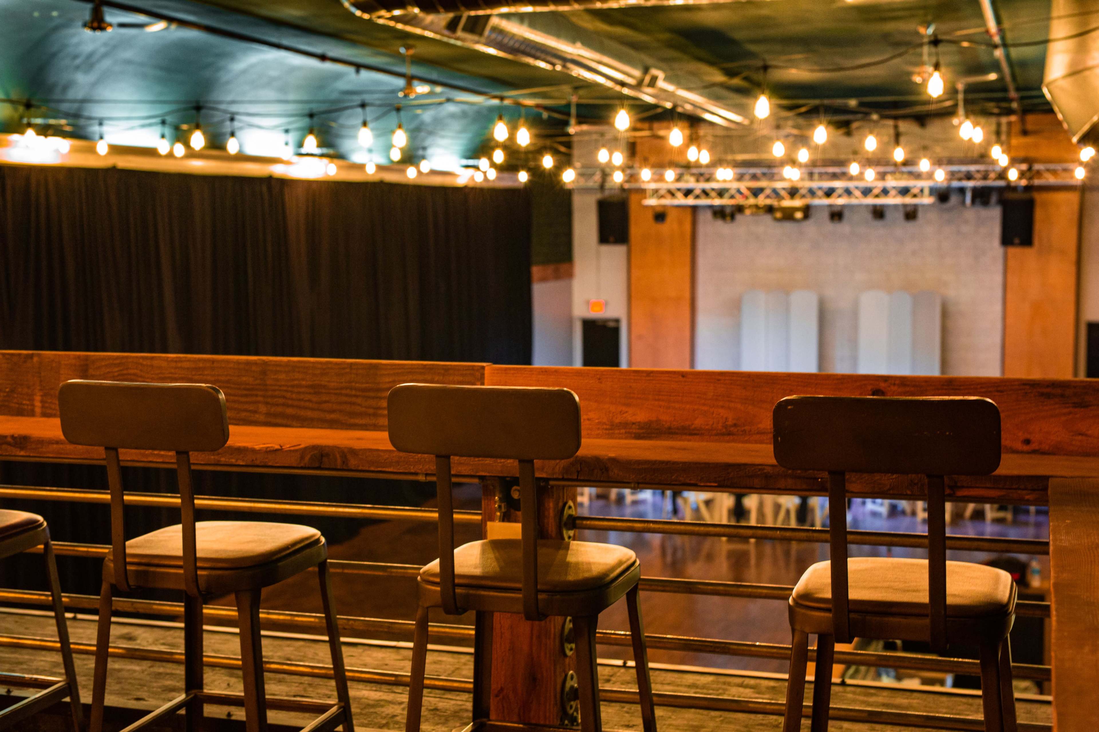 A row of empty wooden stools is positioned along a balcony overlooking a stage area illuminated by hanging lights.