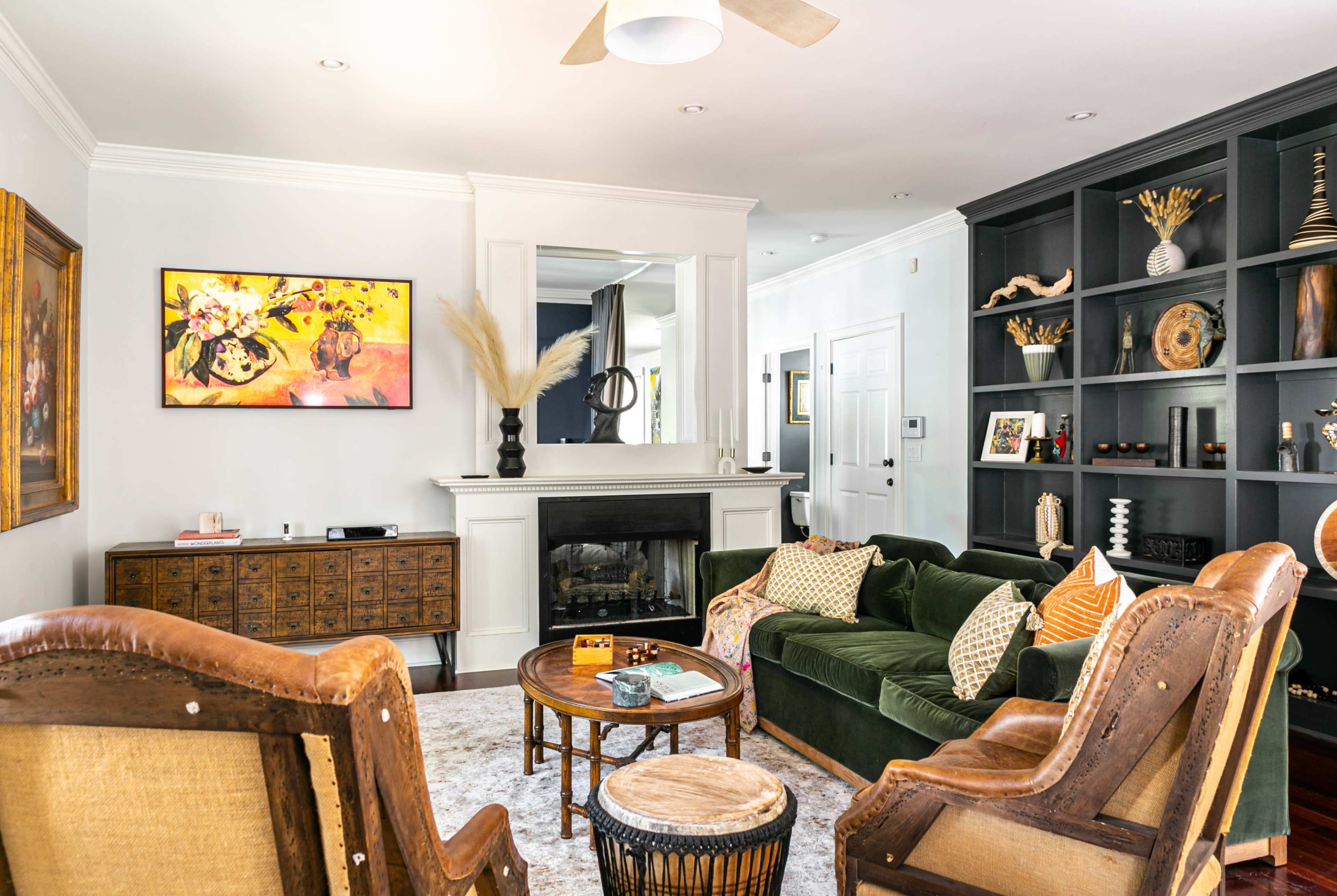 The living room features a green velvet sofa, two wooden armchairs, a round coffee table, and a fireplace, with artwork and decor arranged on the walls and shelves.