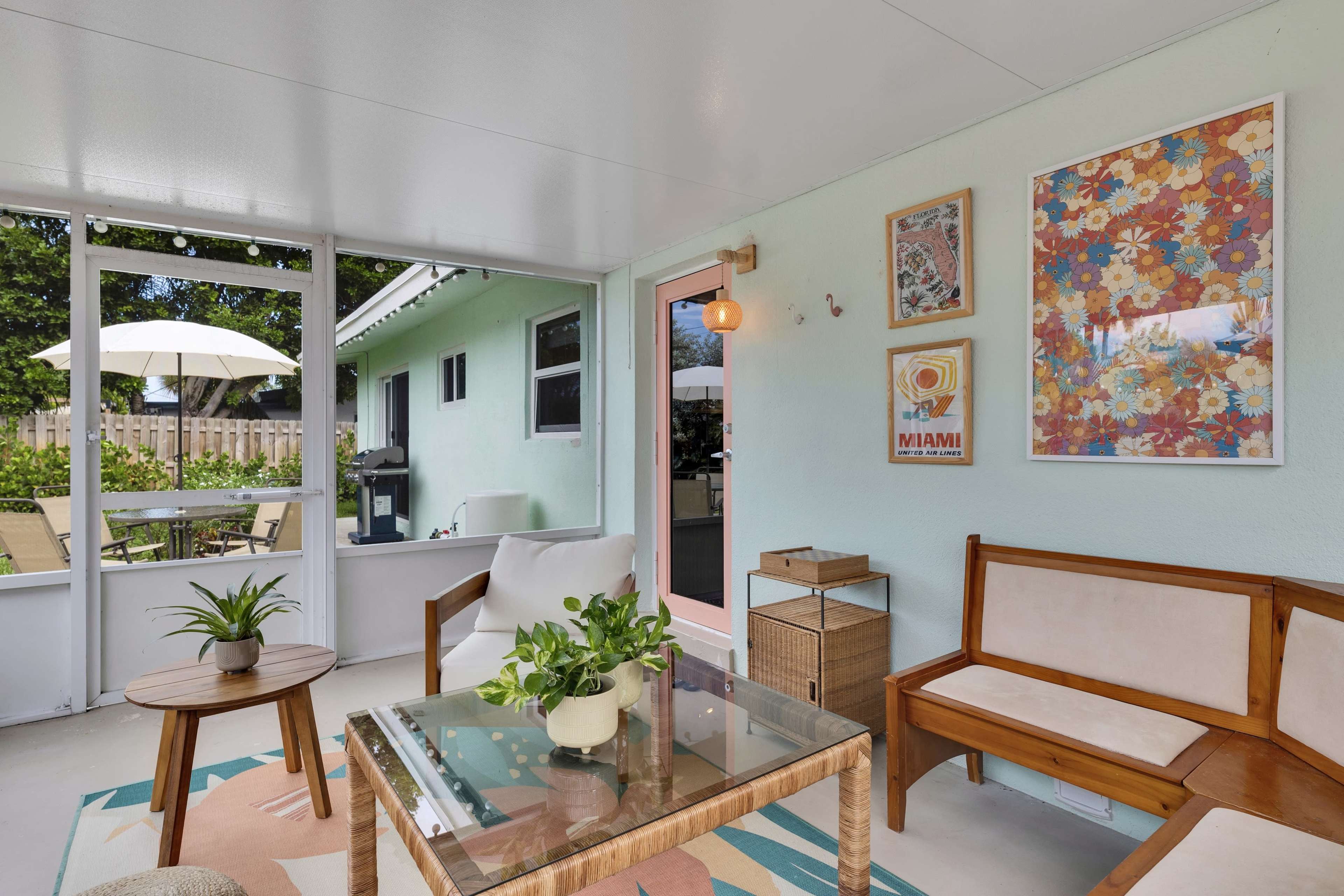 The image shows a sunlit sunroom with a glass table, wooden seating, and colorful artwork on the walls, overlooking a garden through a screened-in porch.