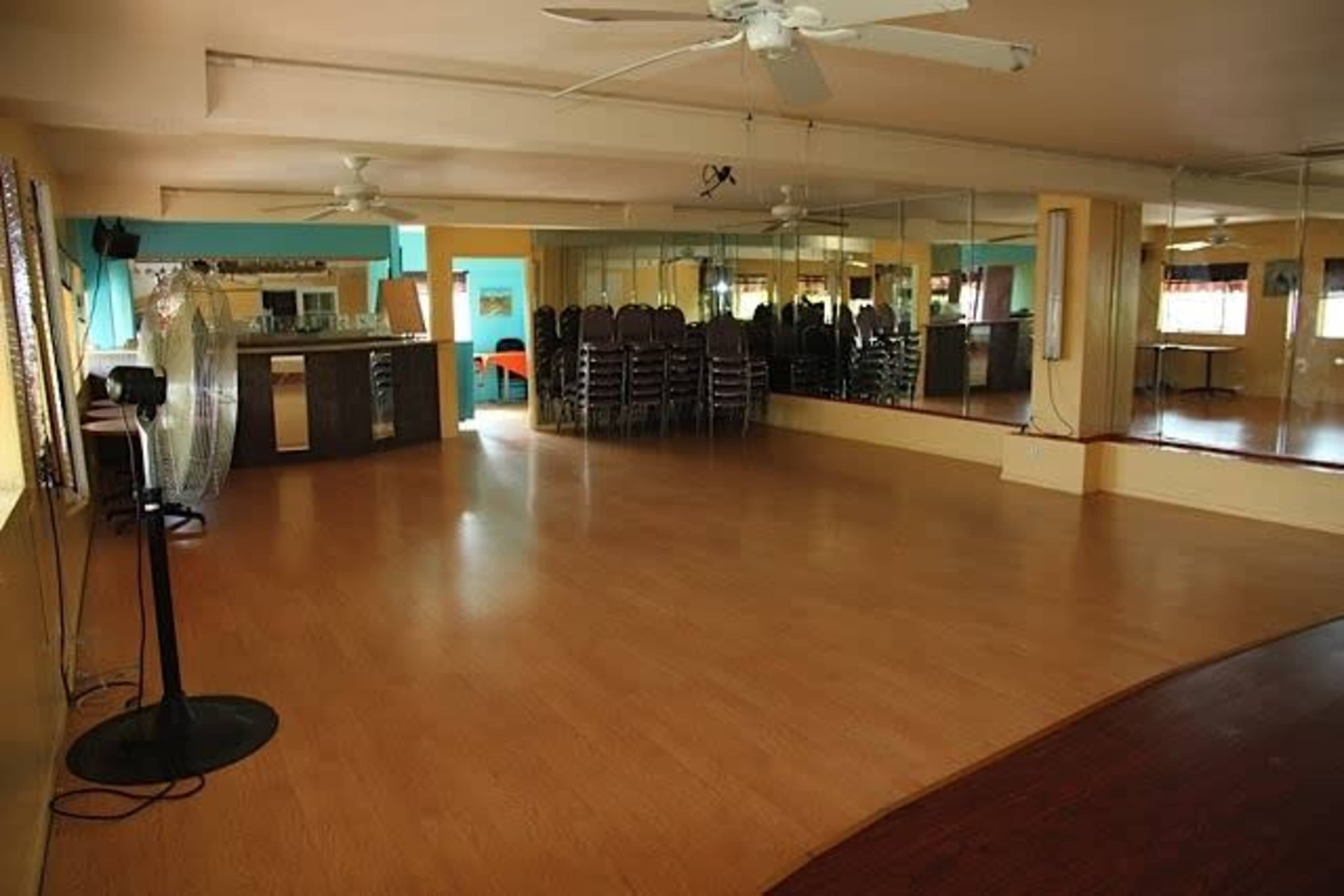 The image shows a spacious dance or activity studio with wooden flooring, mirrors on one wall, and stacked chairs in the background.