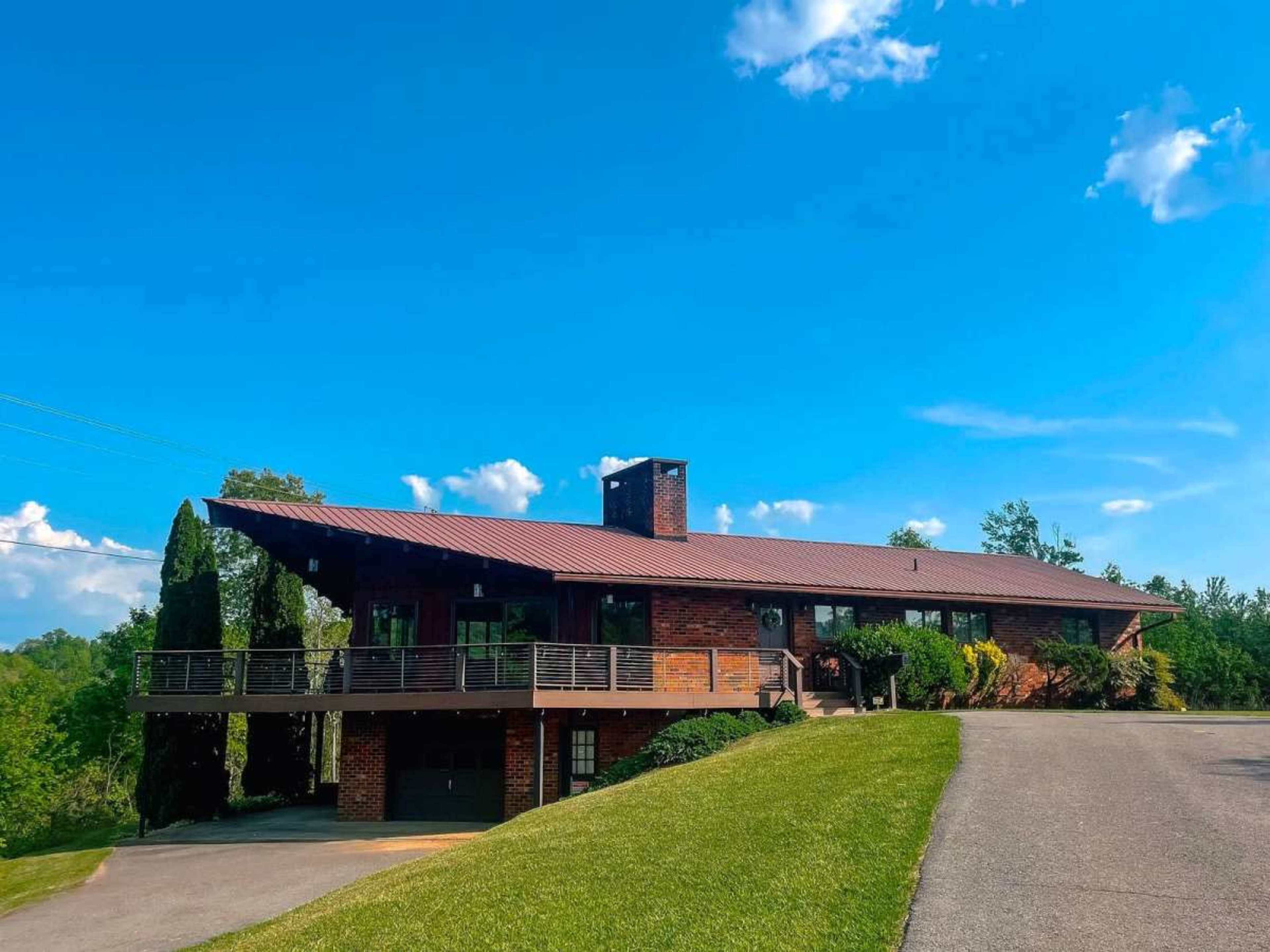 A large, brick house with a metal roof sits atop a green hill under a blue sky.