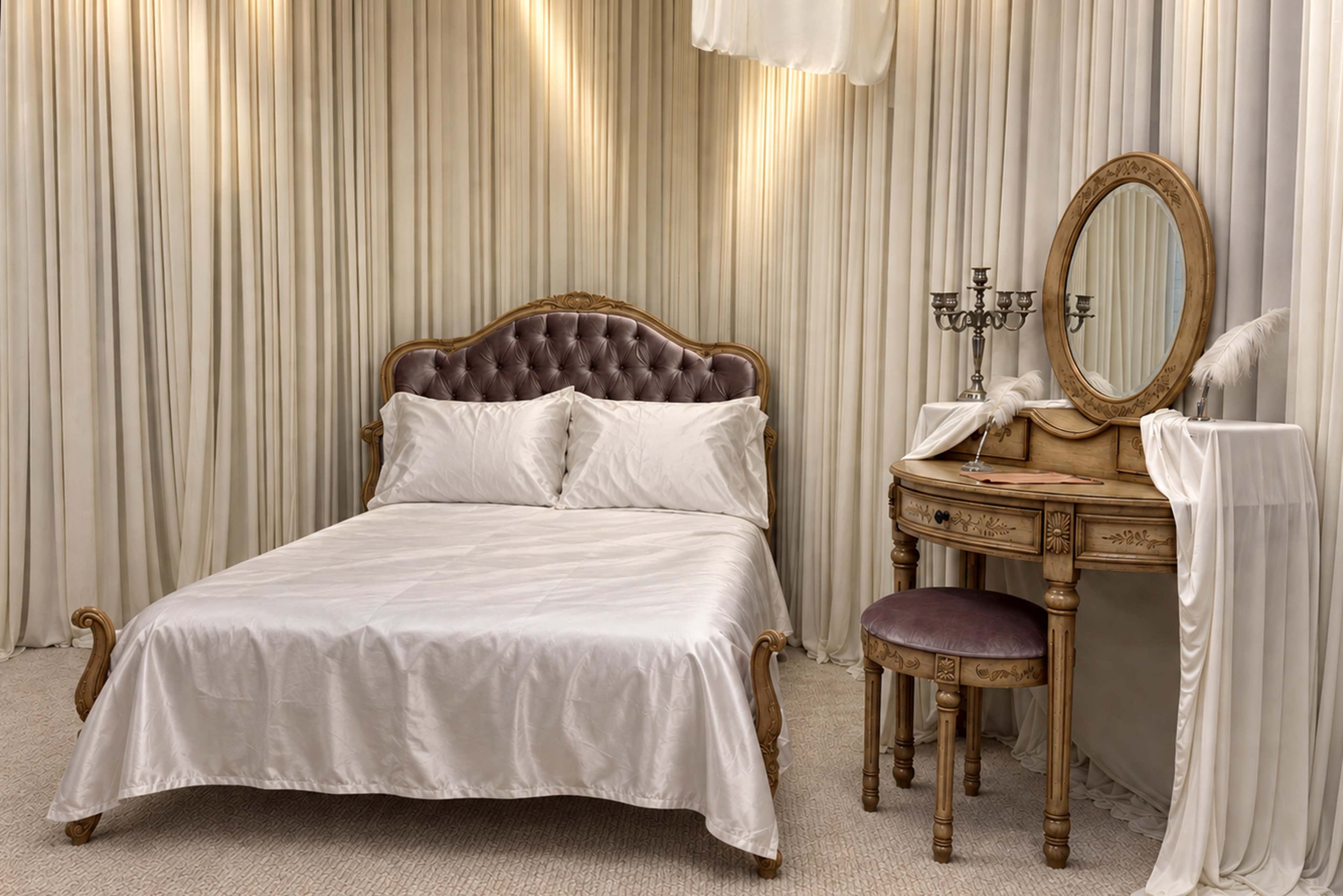 The image depicts a softly lit bedroom featuring an ornate bed with a tufted headboard and a wooden vanity with a round mirror.