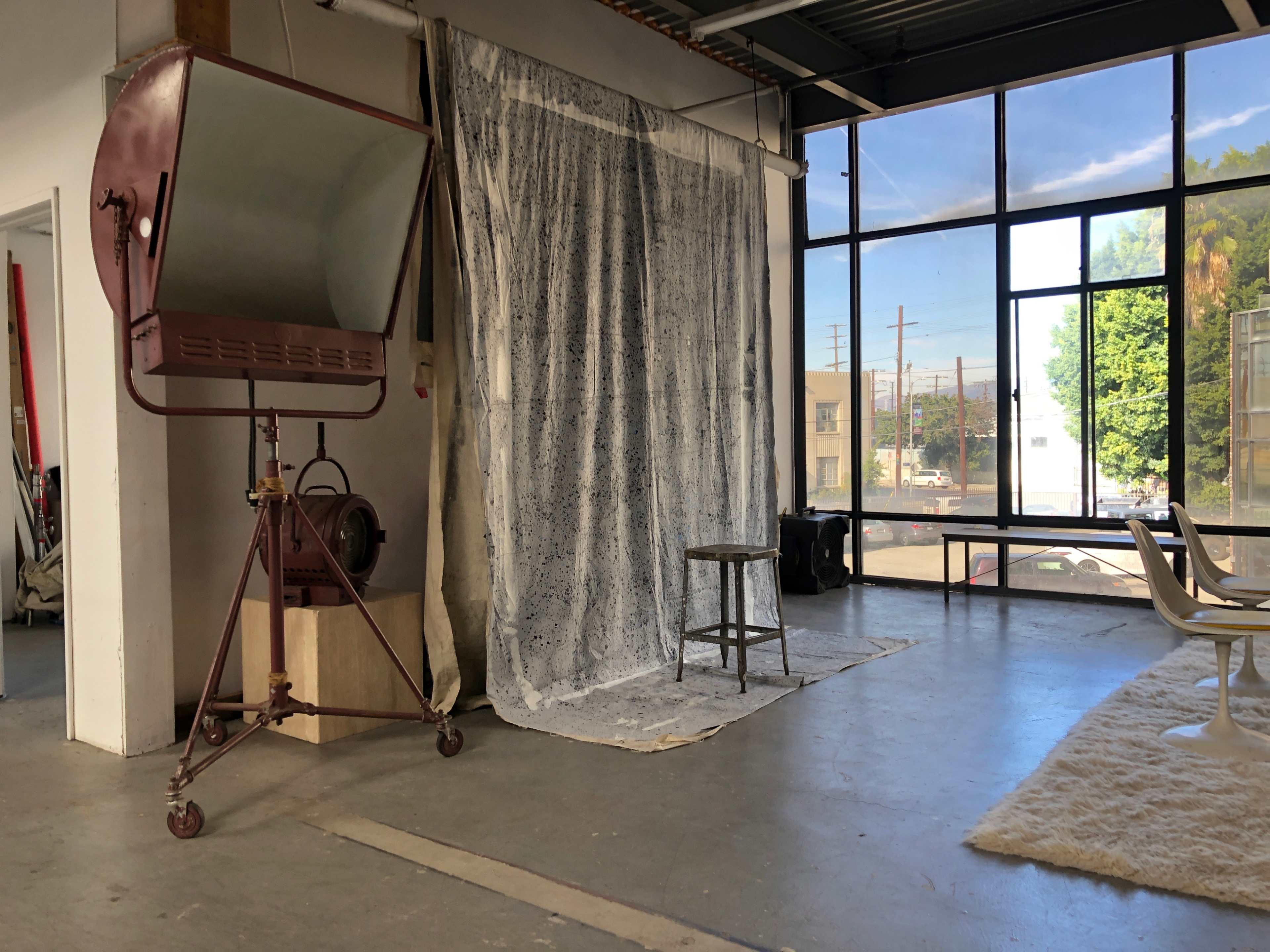 A photography studio with a large light stand, a white backdrop, a stool, and several windows allowing natural light to enter.