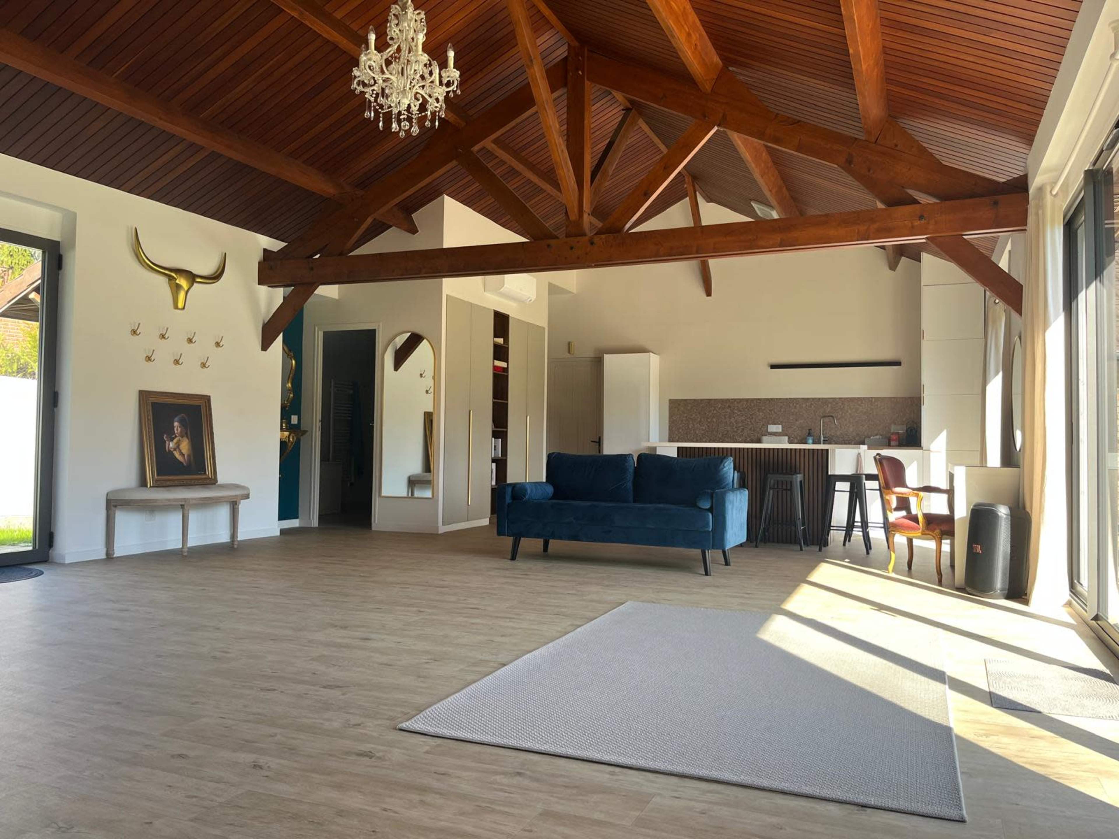 The image shows a spacious, well-lit room with wooden beams, a plush blue couch, and a light area rug on a wooden floor, leading to a modern kitchen area.