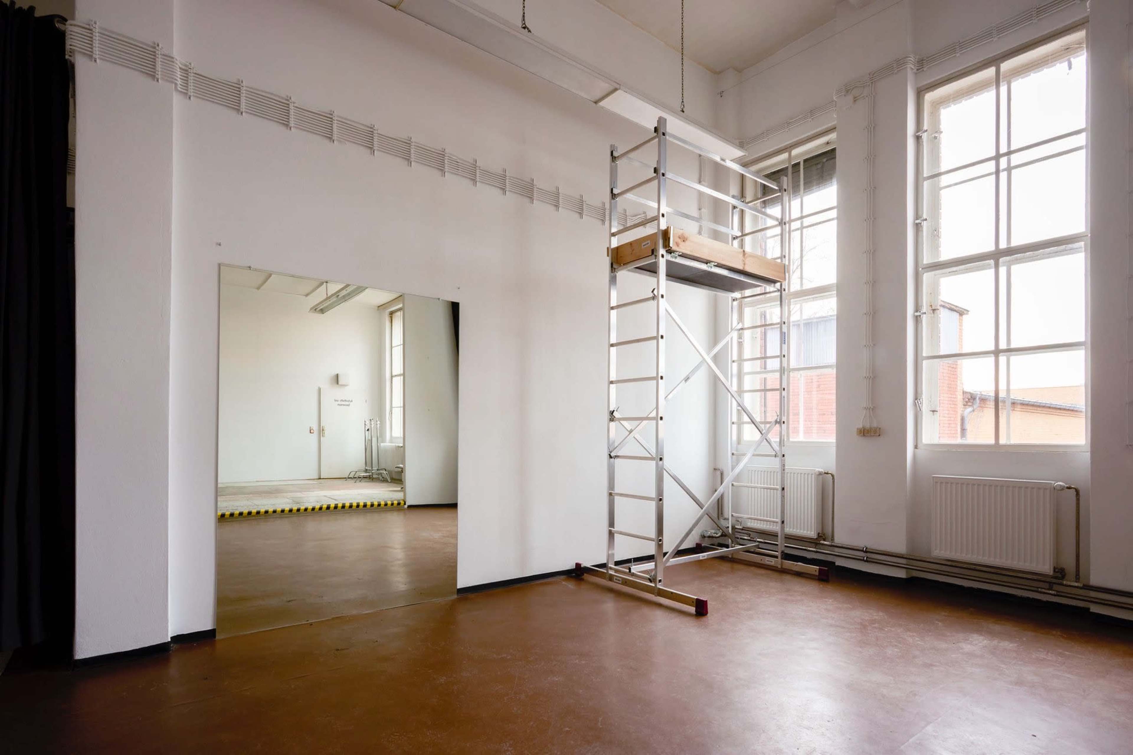 The image shows an empty room with a scaffolding structure and large windows, along with a mirrored wall section.