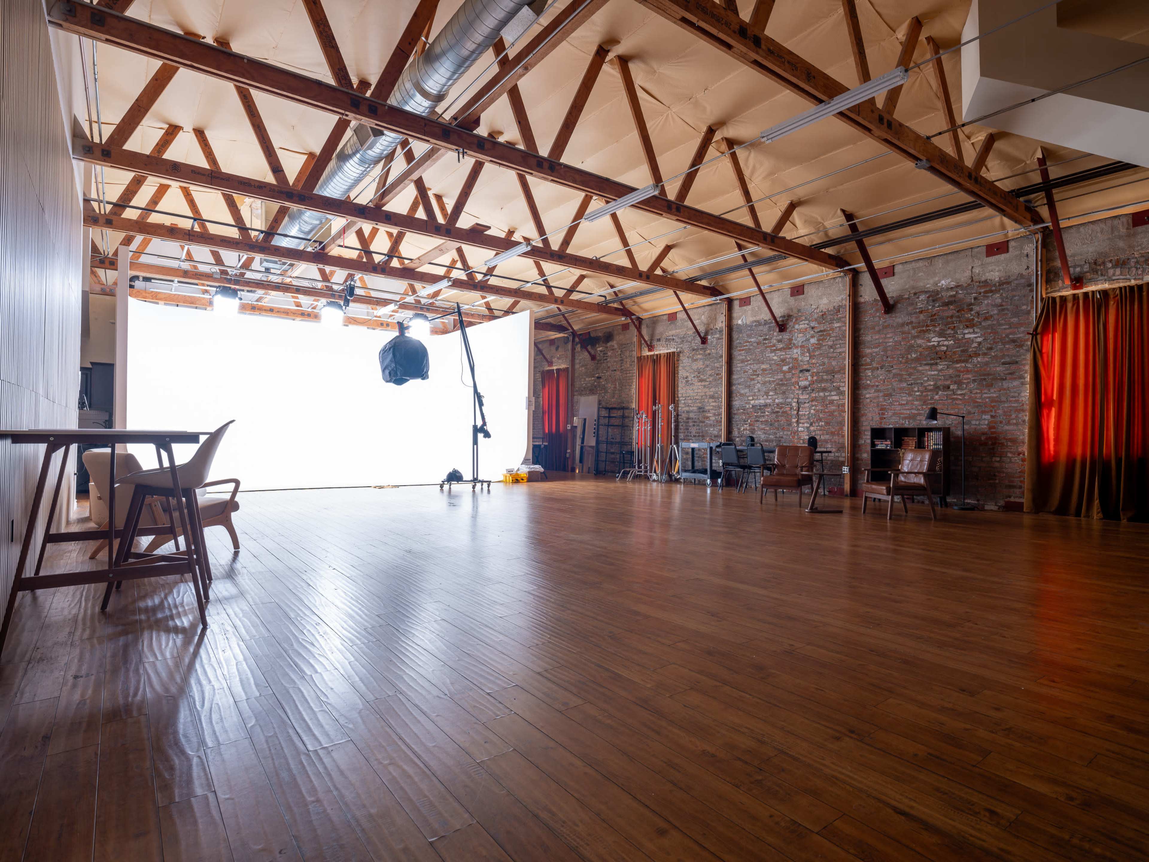 A spacious indoor studio with wooden floors, exposed beams, and various pieces of furniture, including chairs and a table, set against a backdrop of bright light.