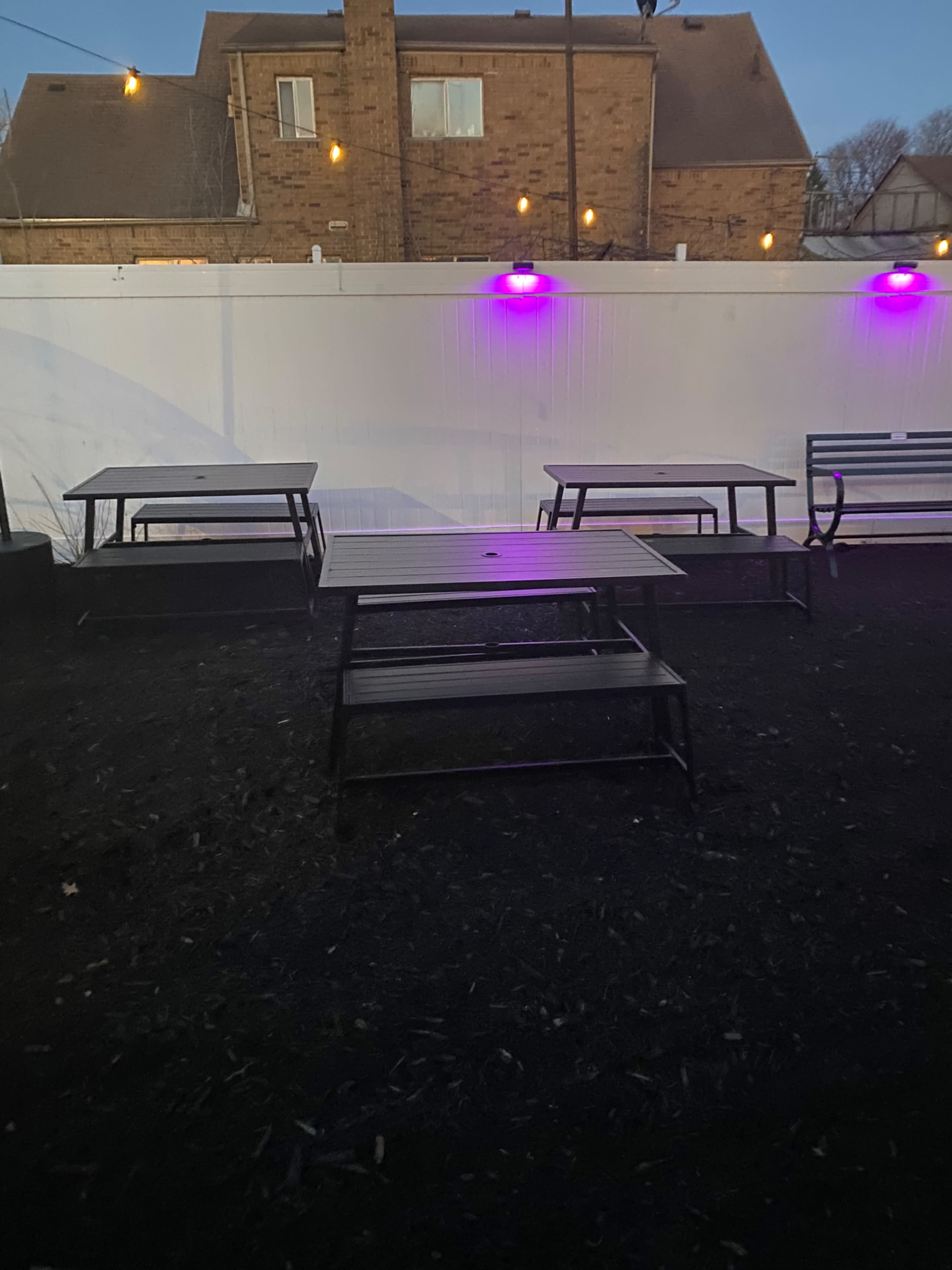 The image shows empty metal tables arranged in an outdoor area near a white fence illuminated by purple lights.