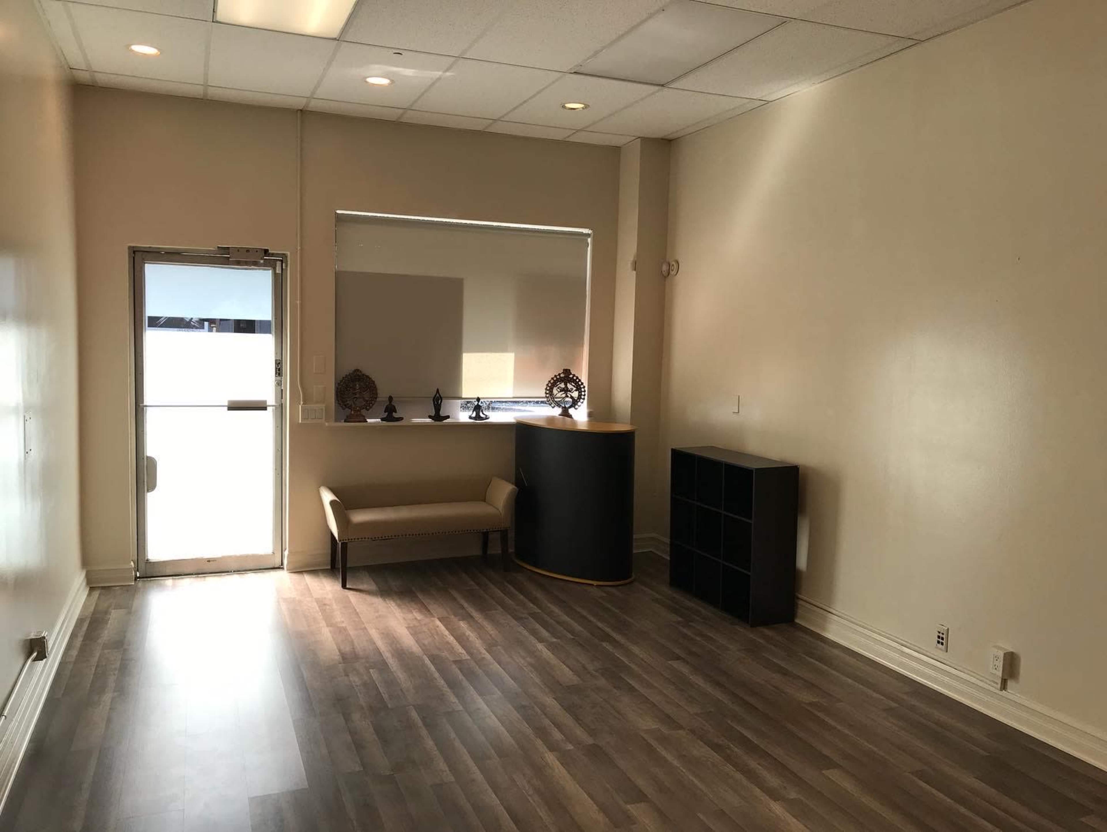 A sparsely furnished interior of a shop or office with light-colored walls, a bench, and a small display area near the entrance.