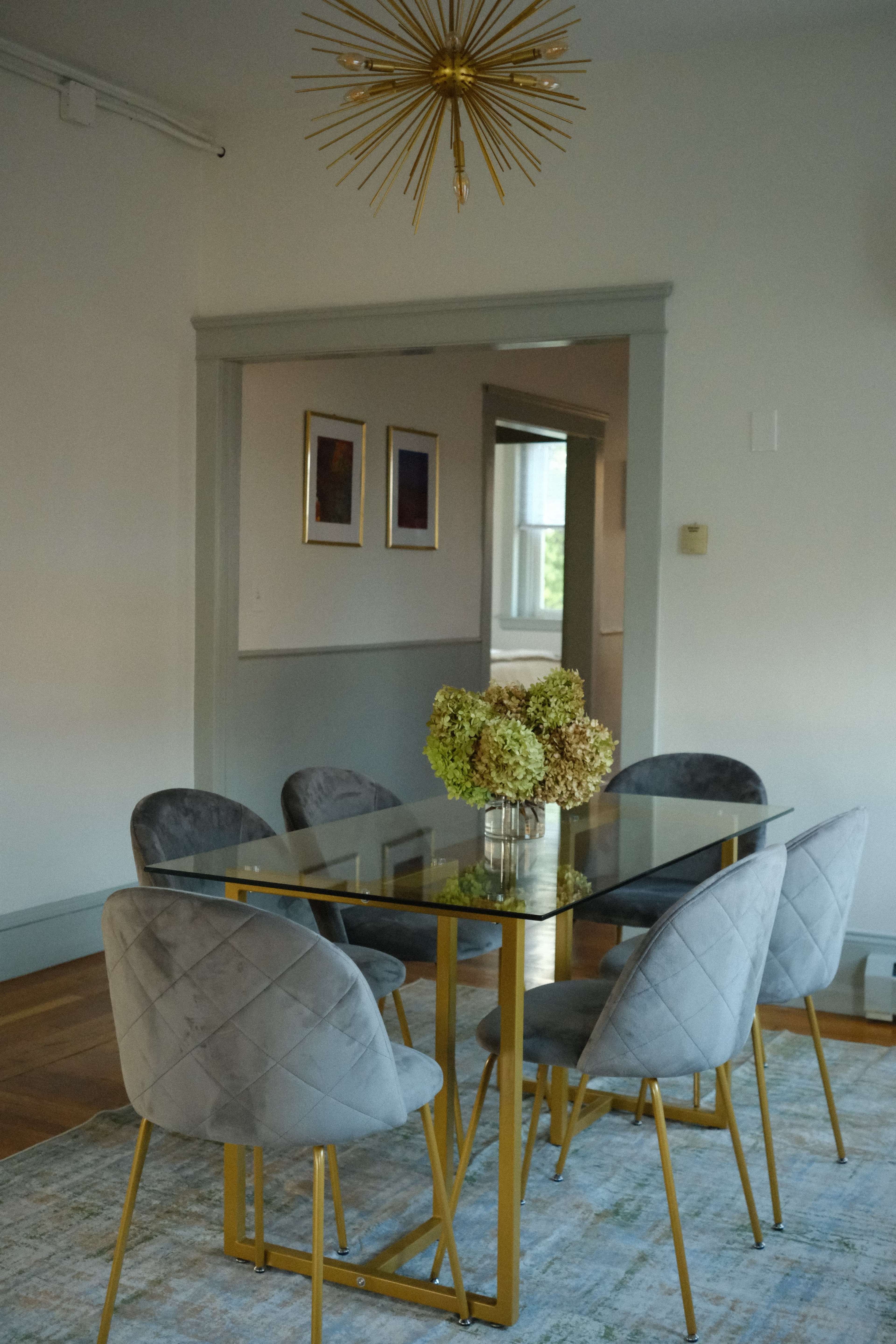 A glass dining table with gold legs is surrounded by four gray chairs and is accentuated by a floral centerpiece, under a starburst chandelier.