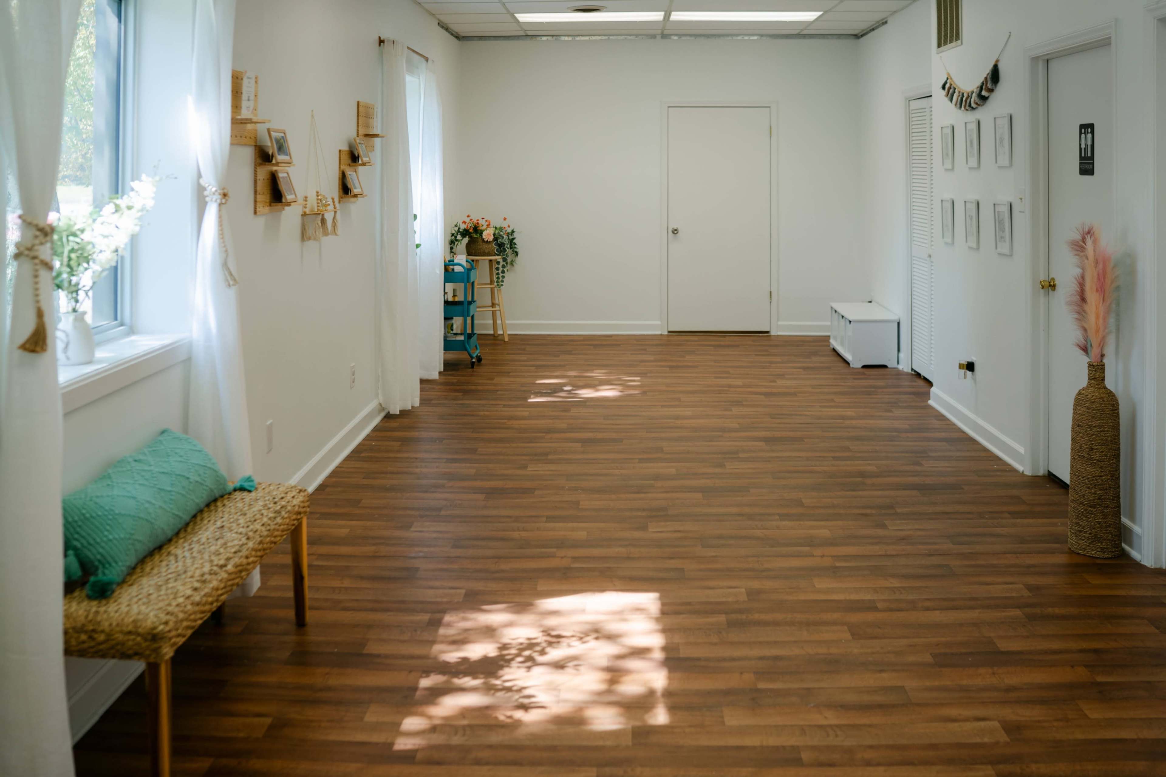 The image depicts a spacious, well-lit room with wooden flooring, adorned with light-colored walls and minimal decorations, including a few plants and framed pictures.